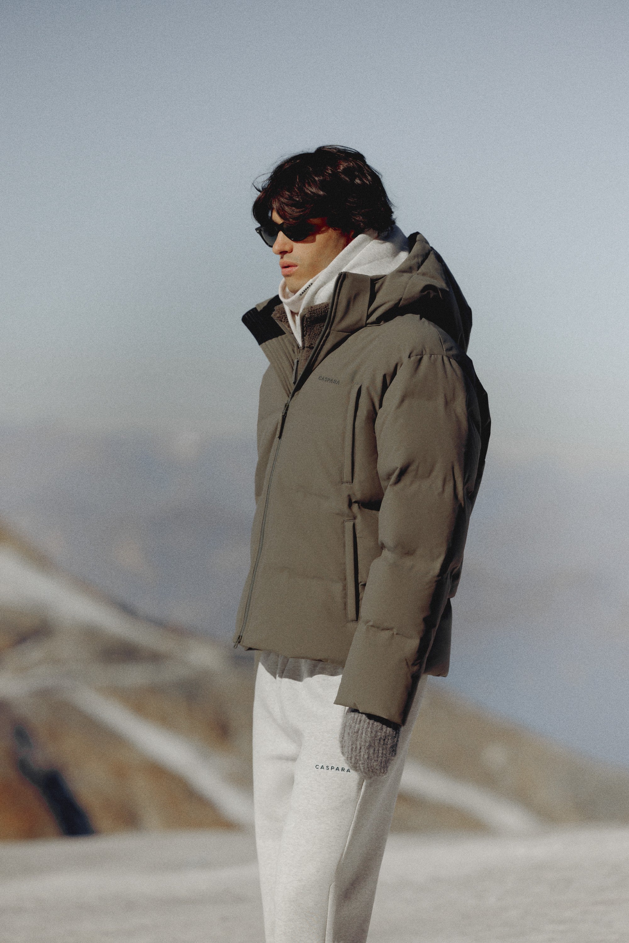 Man wearing a khaki CASPARA Boreal puffer jacket outdoors in the mountains.