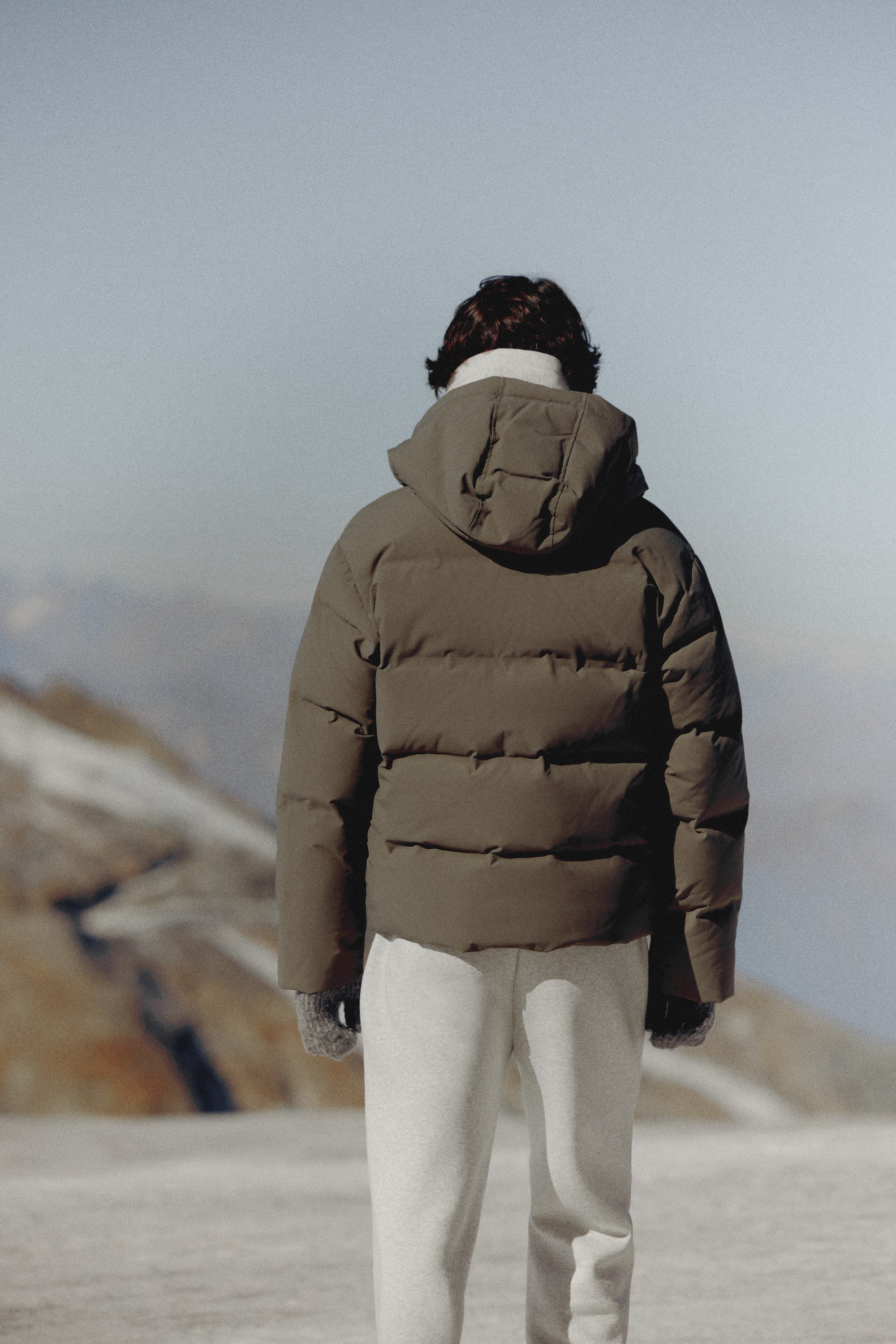 Back view of a man wearing a khaki CASPARA Boreal puffer jacket in the mountains.