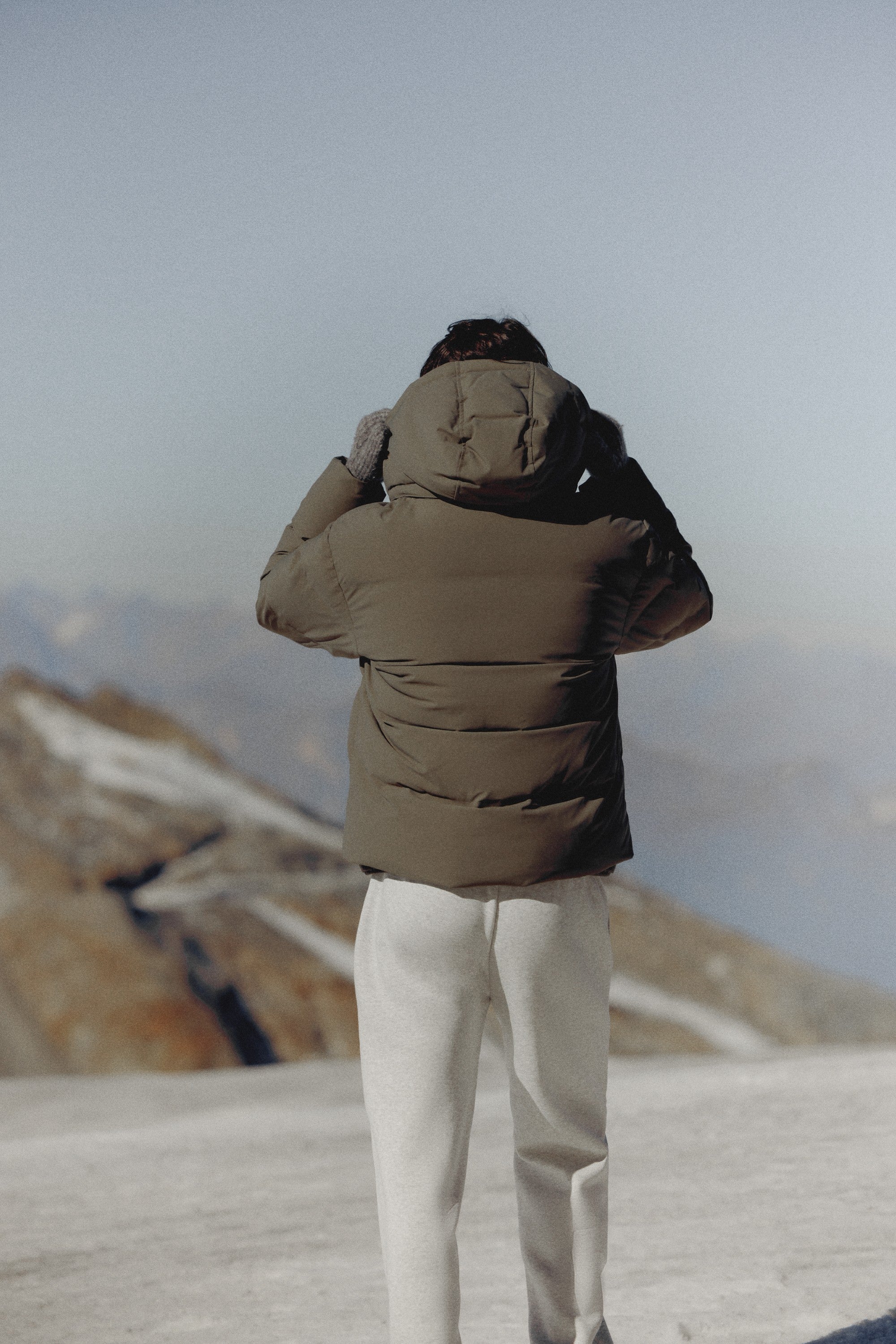 Back view of a man wearing a khaki CASPARA Boreal puffer jacket in a mountain landscape.