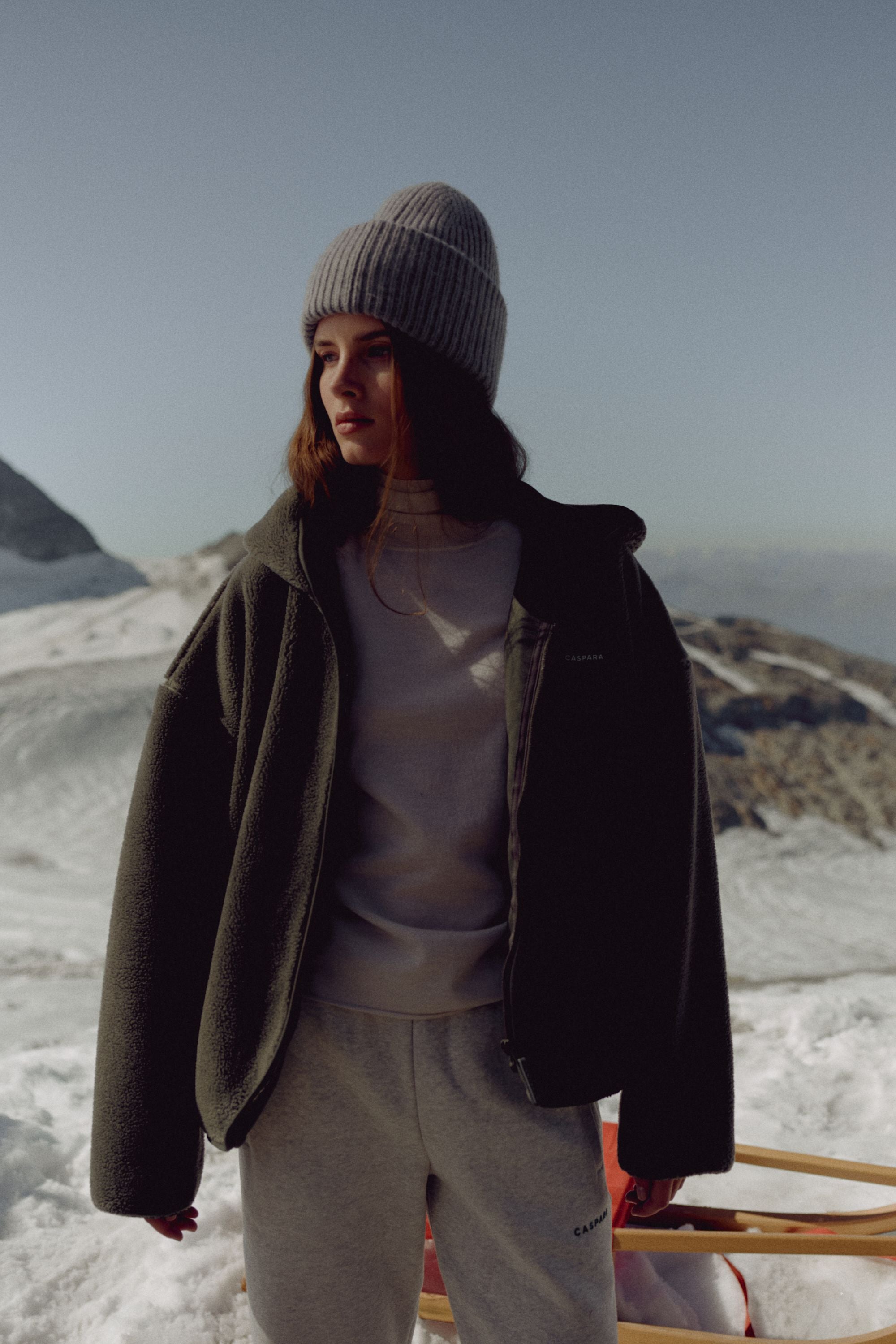 Woman wearing a khaki CASPARA Cloudfleece hooded jacket in a snowy mountain landscape.