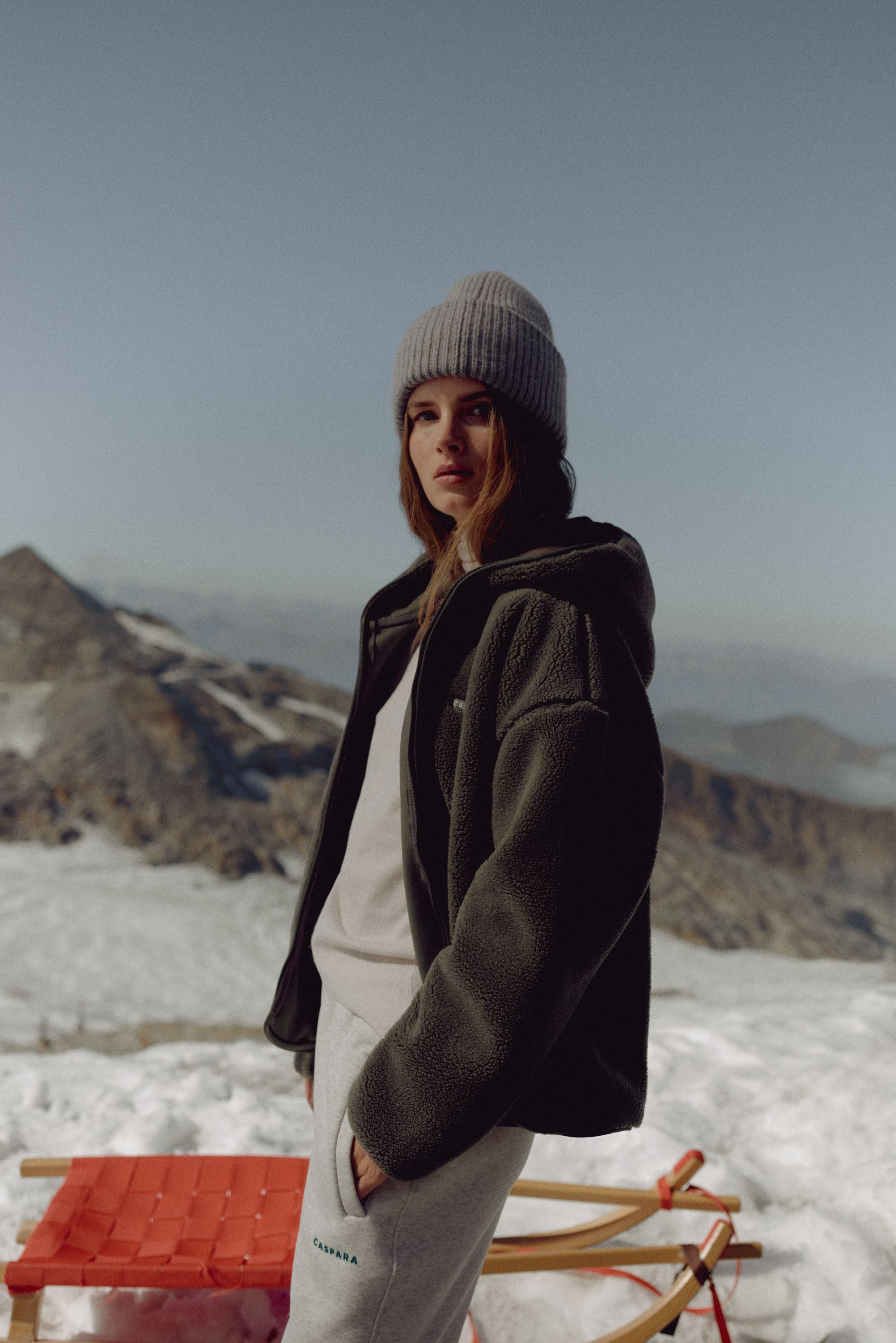 Woman wearing a khaki CASPARA Cloudfleece hooded jacket against a snowy mountain backdrop.