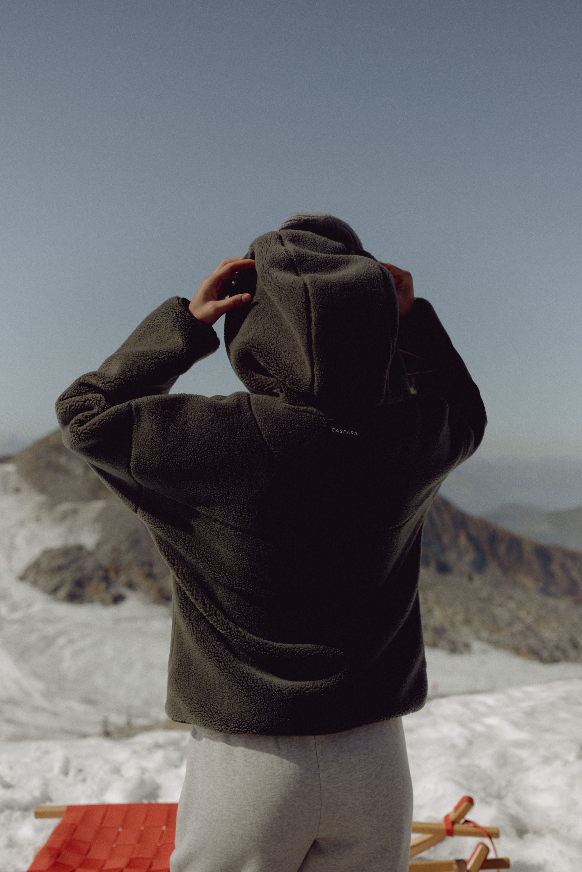 Woman wearing a khaki CASPARA Cloudfleece hooded jacket in a snowy mountain landscape.