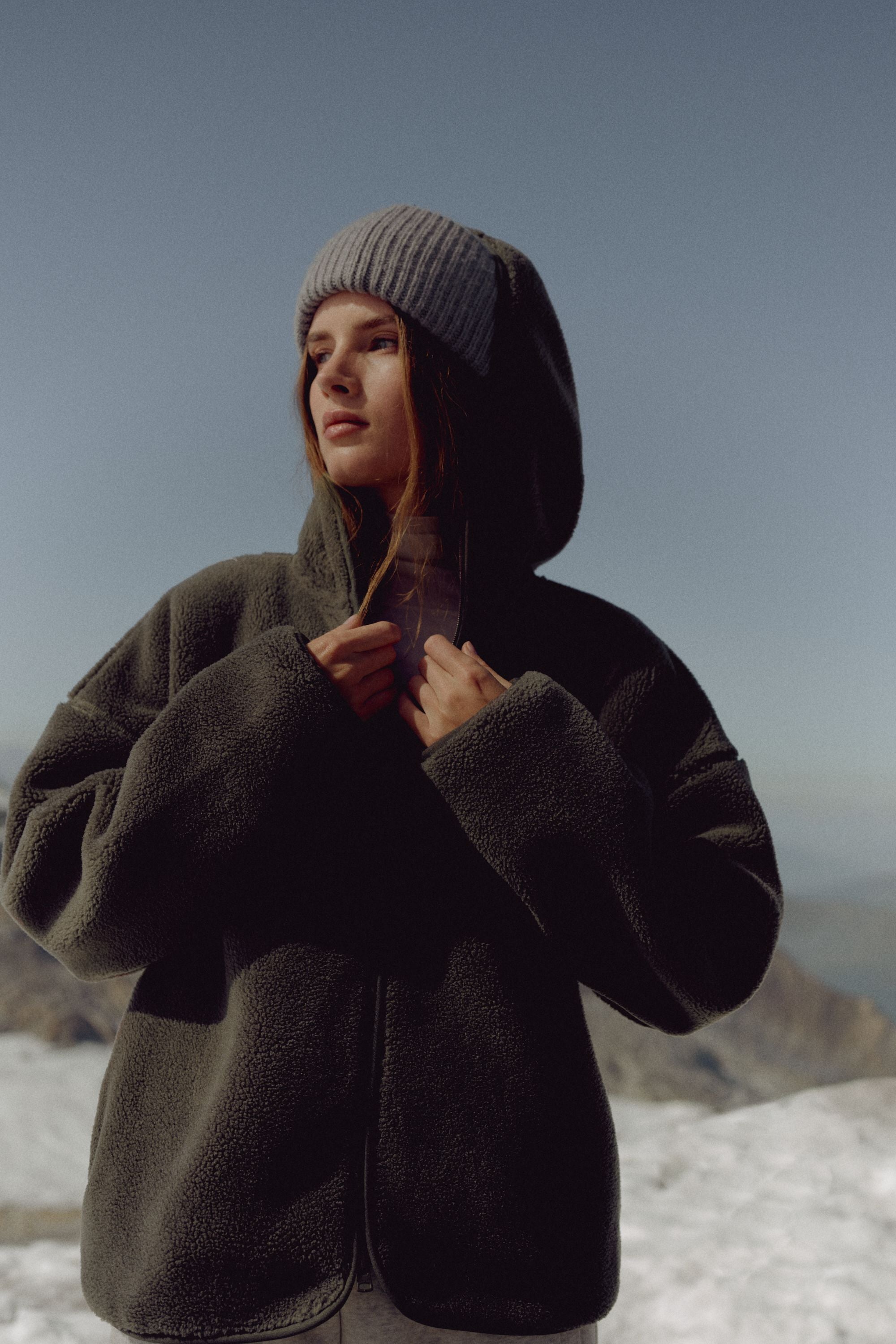 Woman wearing a khaki CASPARA Cloudfleece hooded jacket in a snowy mountain landscape.