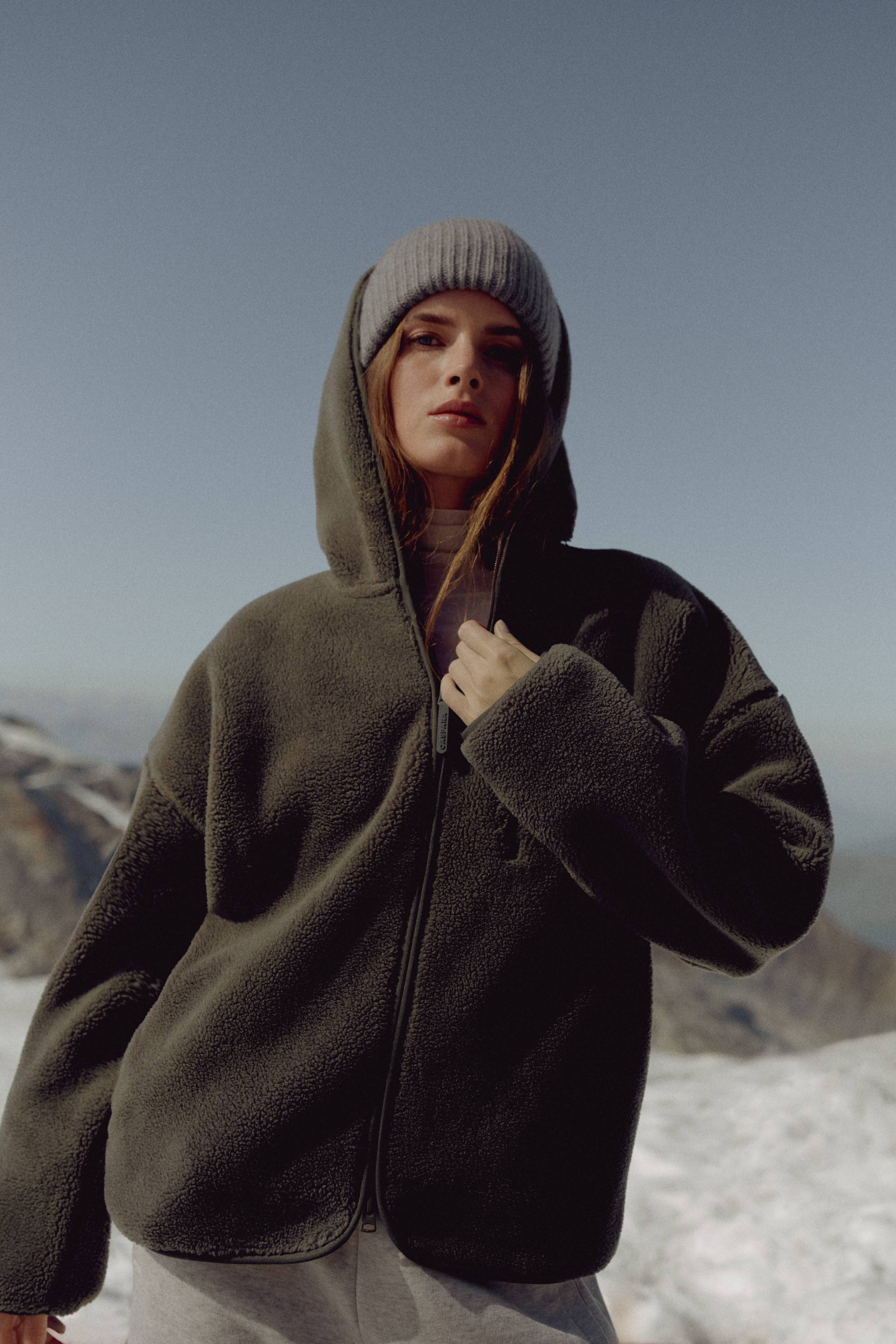 Woman wearing a khaki CASPARA Cloudfleece hooded jacket in a mountain landscape.
