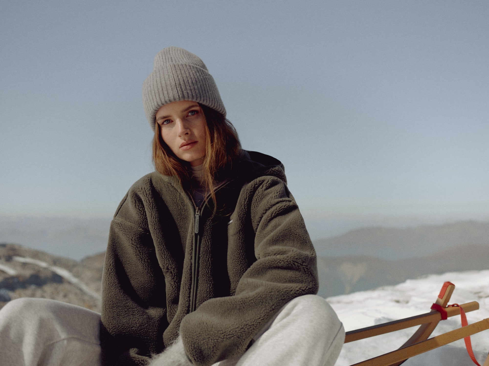 Woman wearing a khaki CASPARA Cloudfleece hooded jacket in a snowy mountain setting.