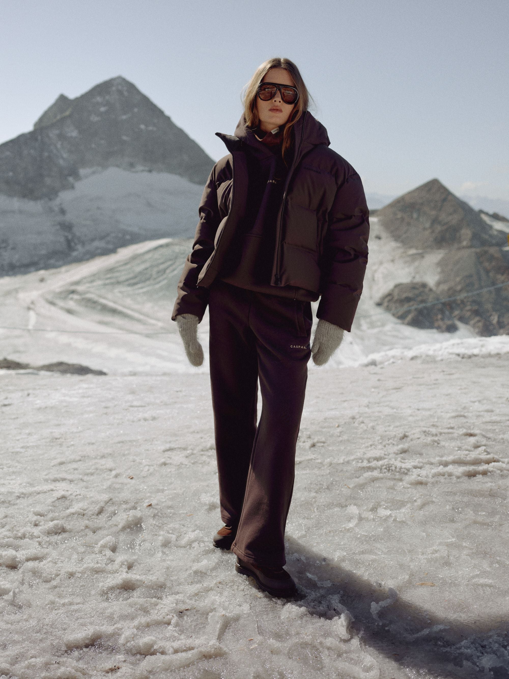 Woman wearing a cocoa bean CASPARA Aurora puffer jacket in a snowy mountain setting.