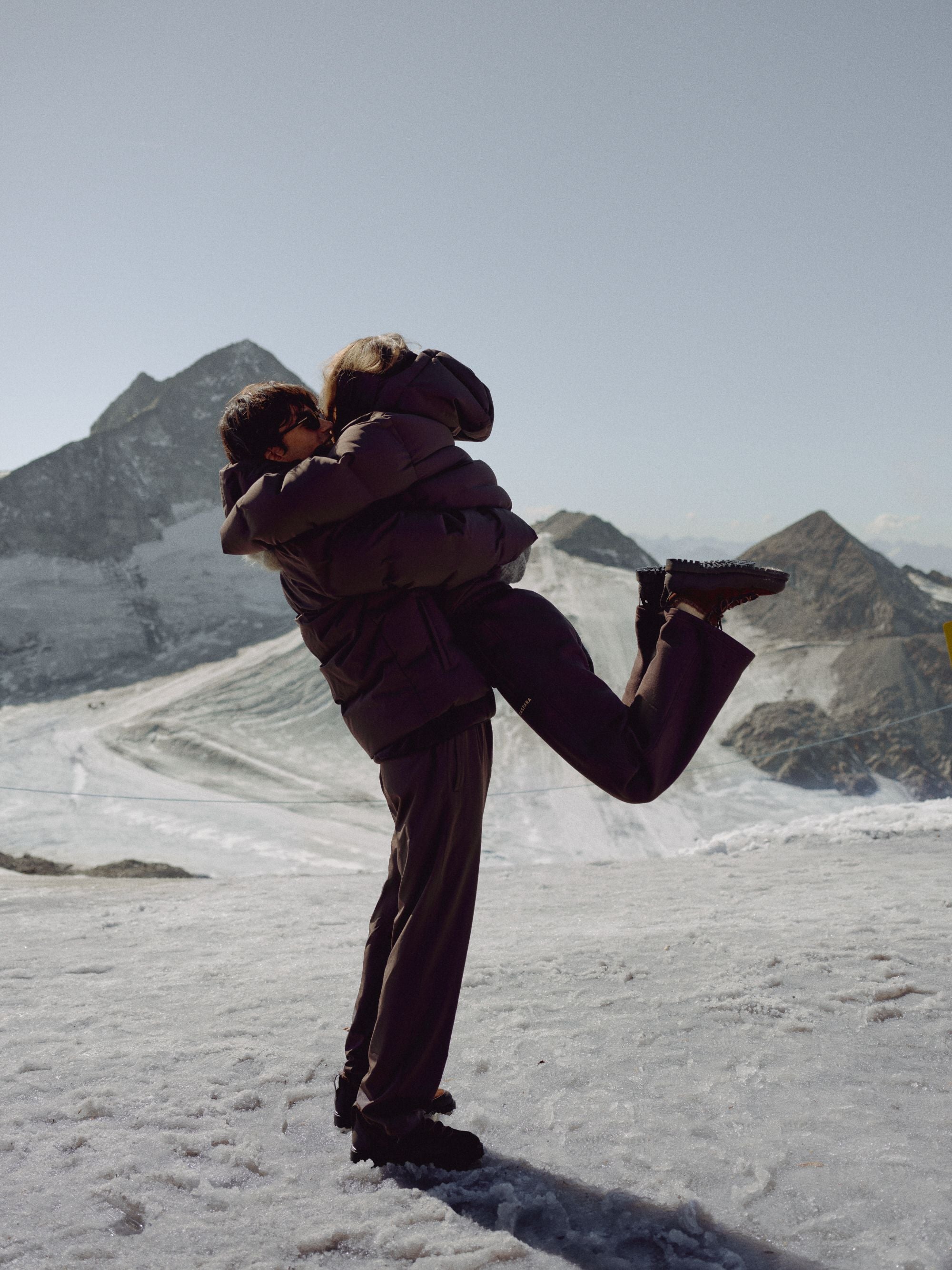 Man lifting a woman wearing a cocoa bean CASPARA Aurora puffer jacket on a snowy mountain.