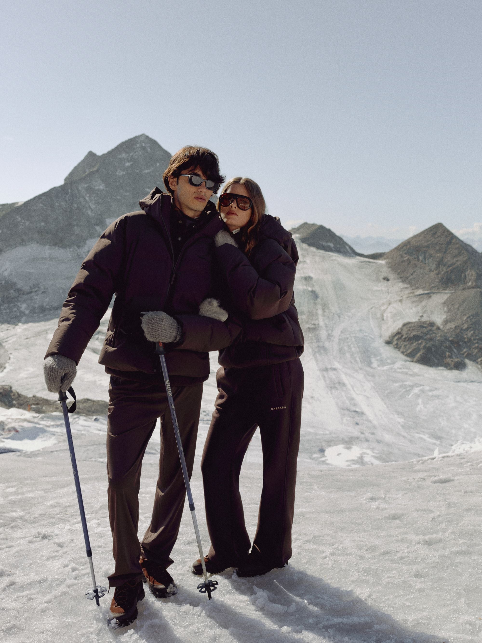 Man and woman wearing cocoa bean CASPARA puffer jackets on a snowy mountain slope.