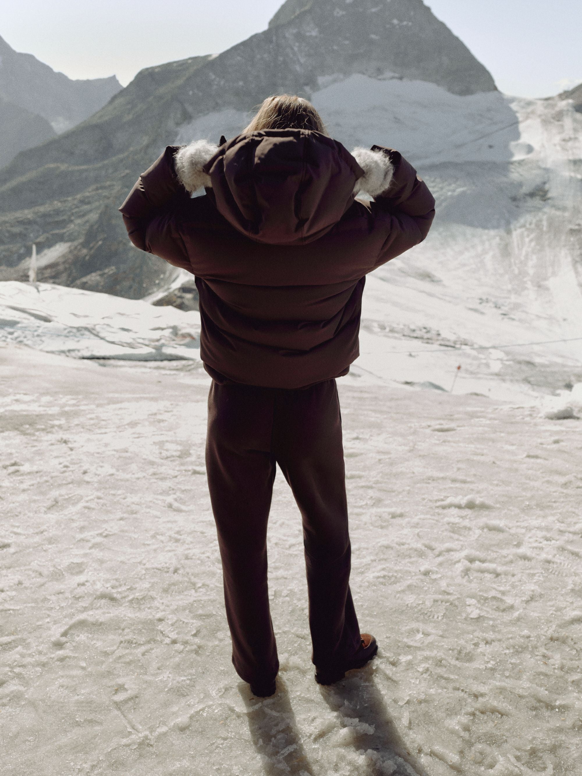 Back view of a woman wearing a cocoa bean CASPARA Aurora puffer jacket in snowy mountains.