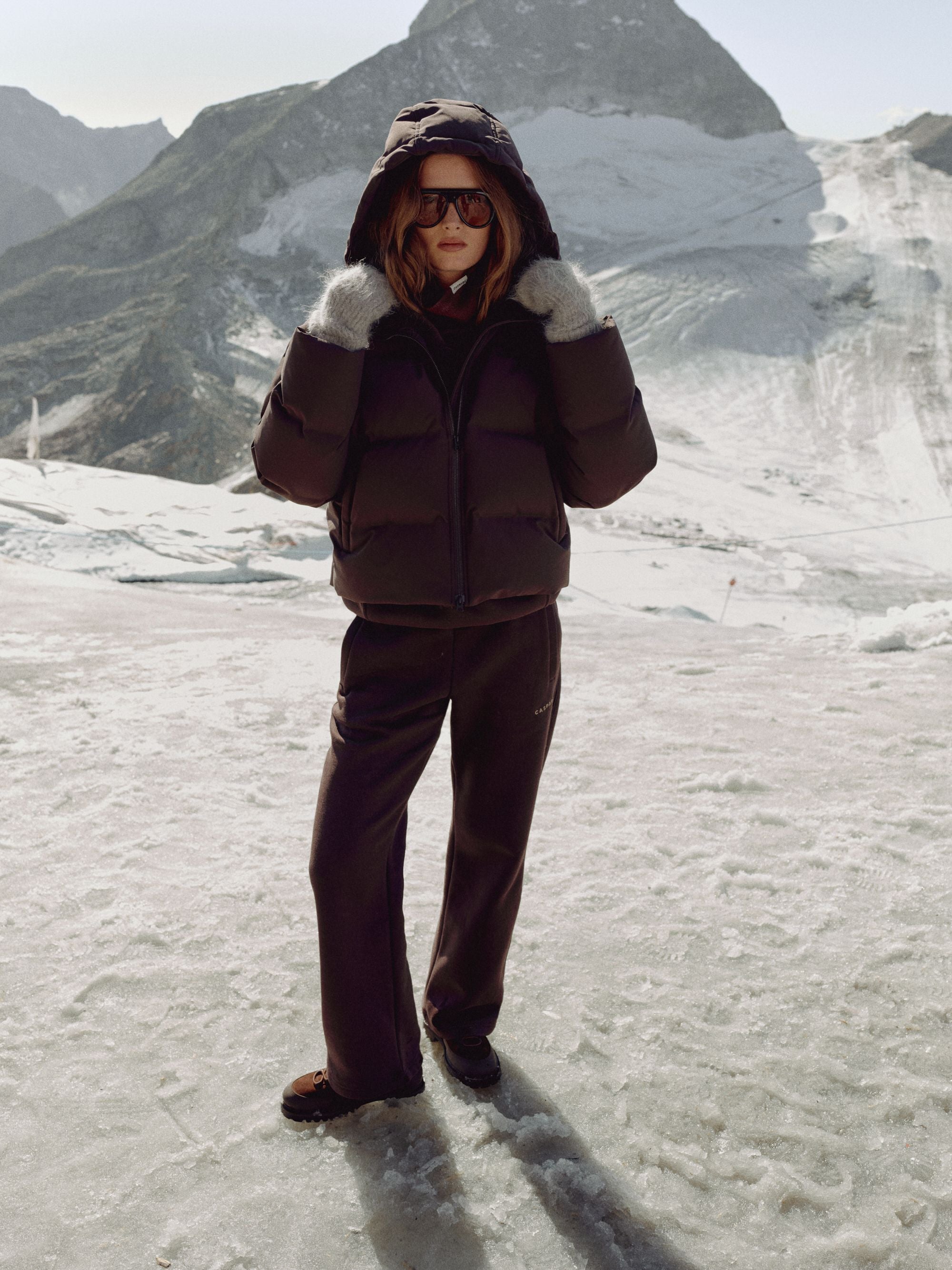 Woman wearing a cocoa bean CASPARA Aurora puffer jacket in a snowy mountain landscape.