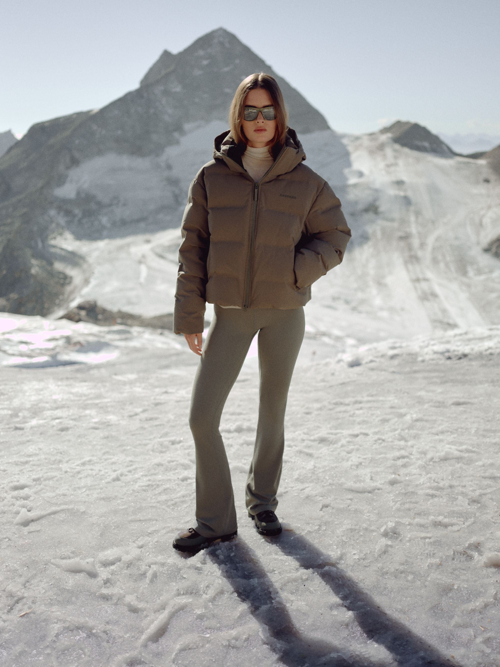 Woman wearing a khaki CASPARA Aurora puffer jacket in a snowy mountain landscape.