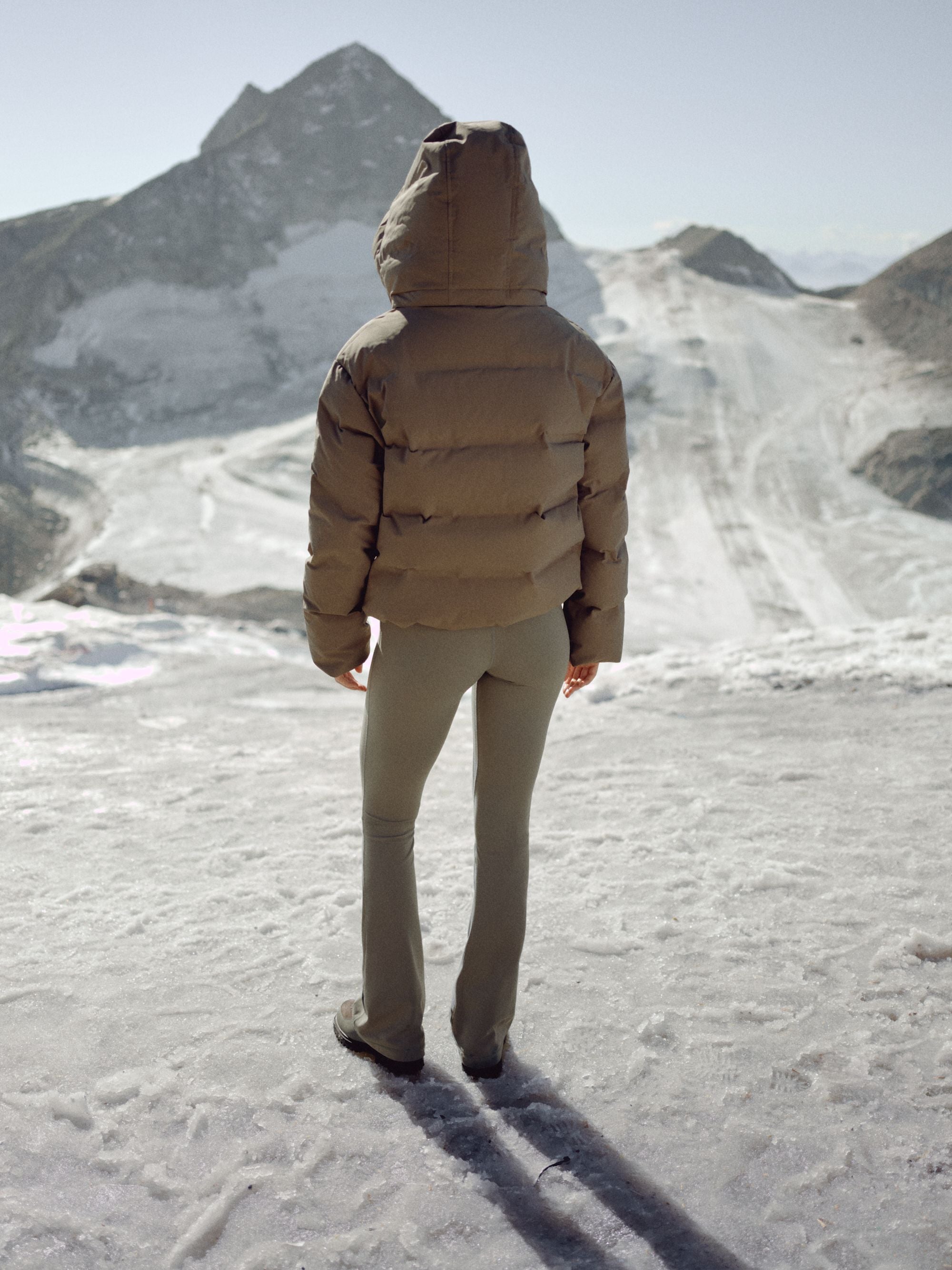 Back view of a woman wearing a khaki CASPARA Aurora puffer jacket in a snowy mountain landscape.