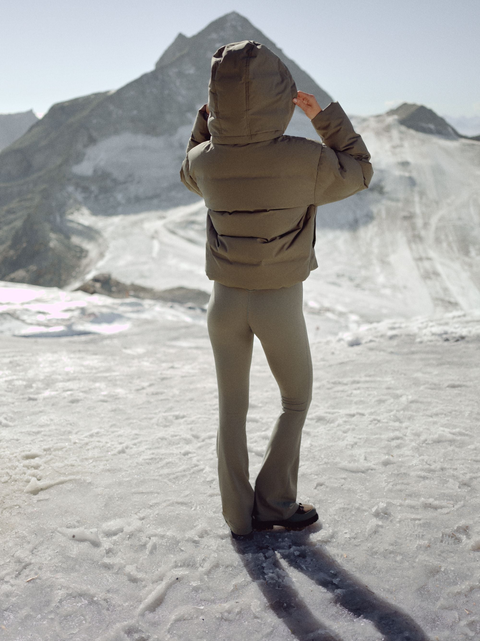 Woman seen from behind wearing a khaki CASPARA Aurora puffer jacket in a snowy mountain landscape.