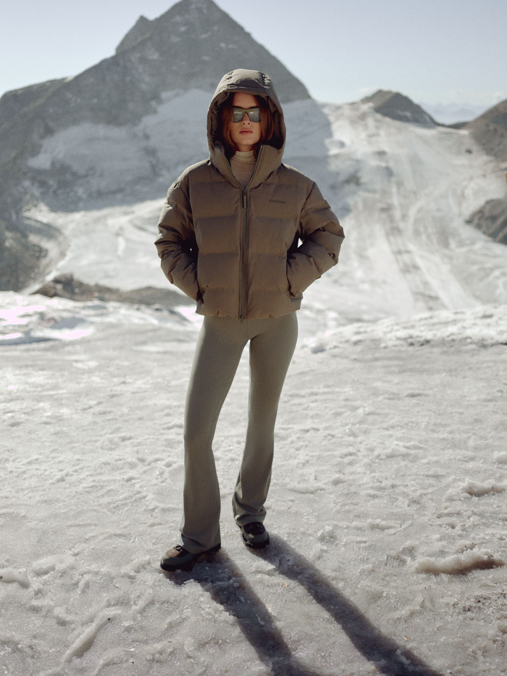 Woman wearing a khaki CASPARA Aurora puffer jacket in a snowy mountain landscape.