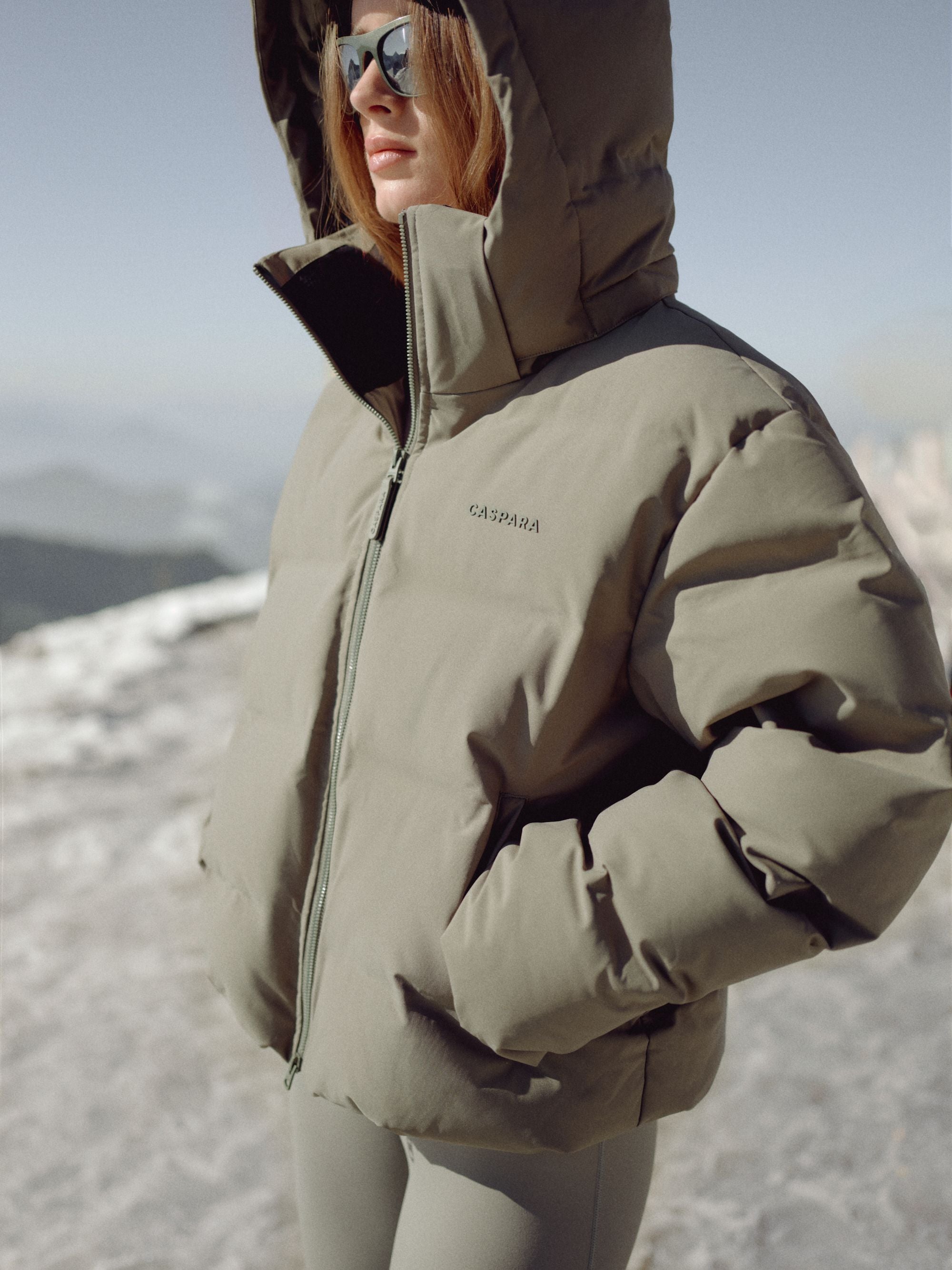 Woman wearing a khaki CASPARA Aurora puffer jacket with the hood up in a snowy mountain landscape.
