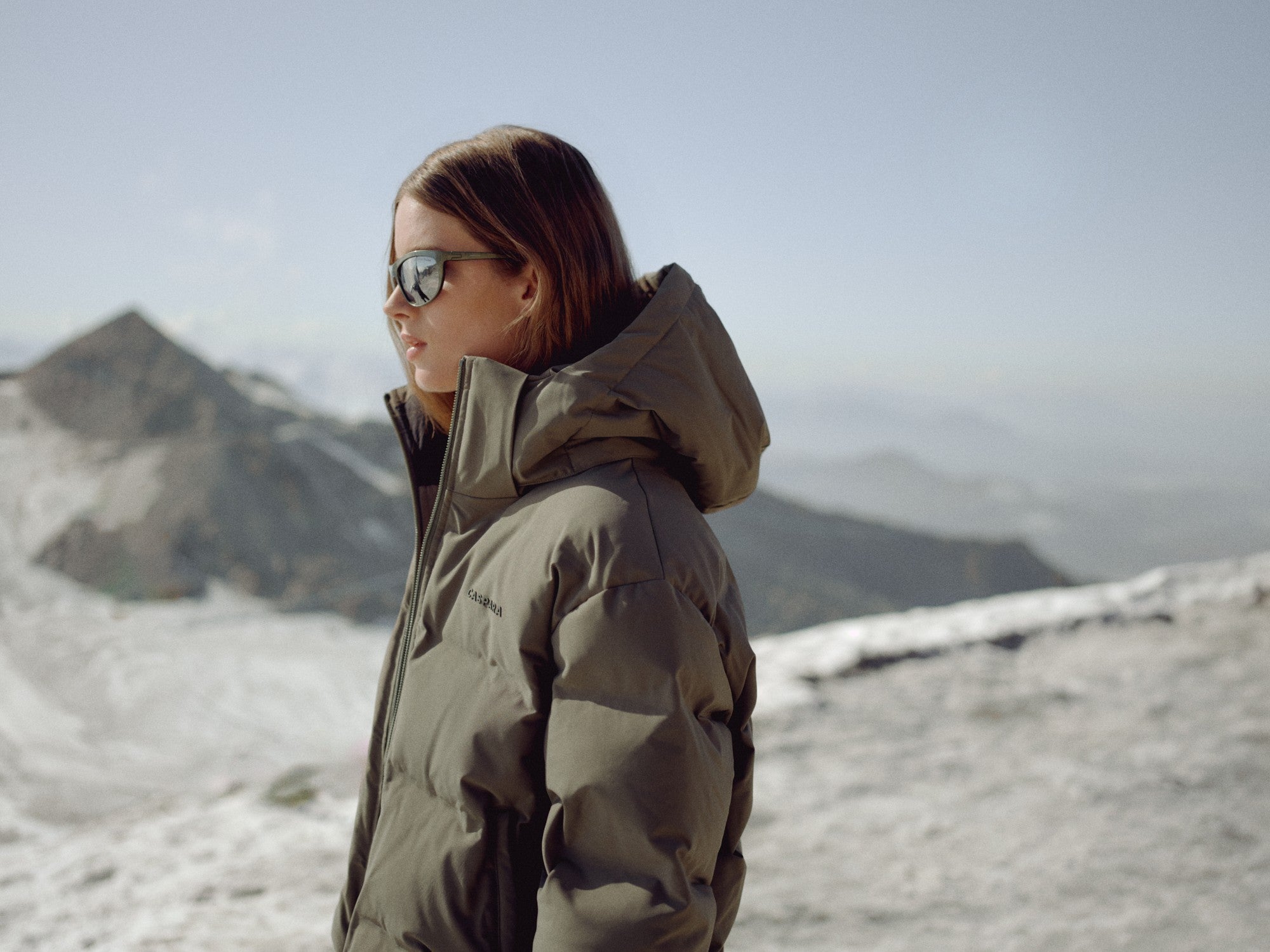 Woman wearing a khaki CASPARA Aurora puffer jacket in a snowy mountain landscape.