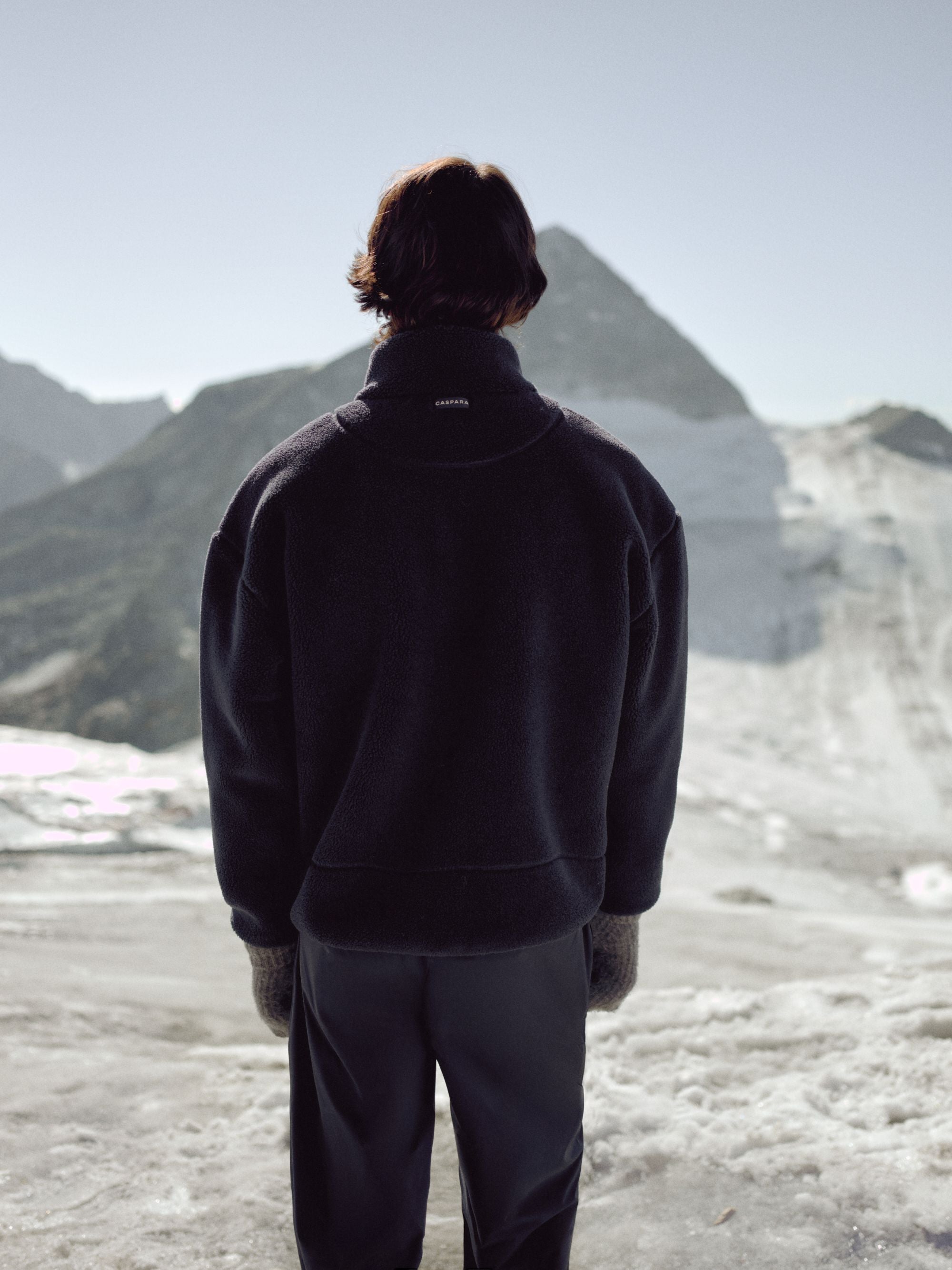 Back view of a man wearing a navy CASPARA Cloudfleece jacket in a snowy mountain landscape.