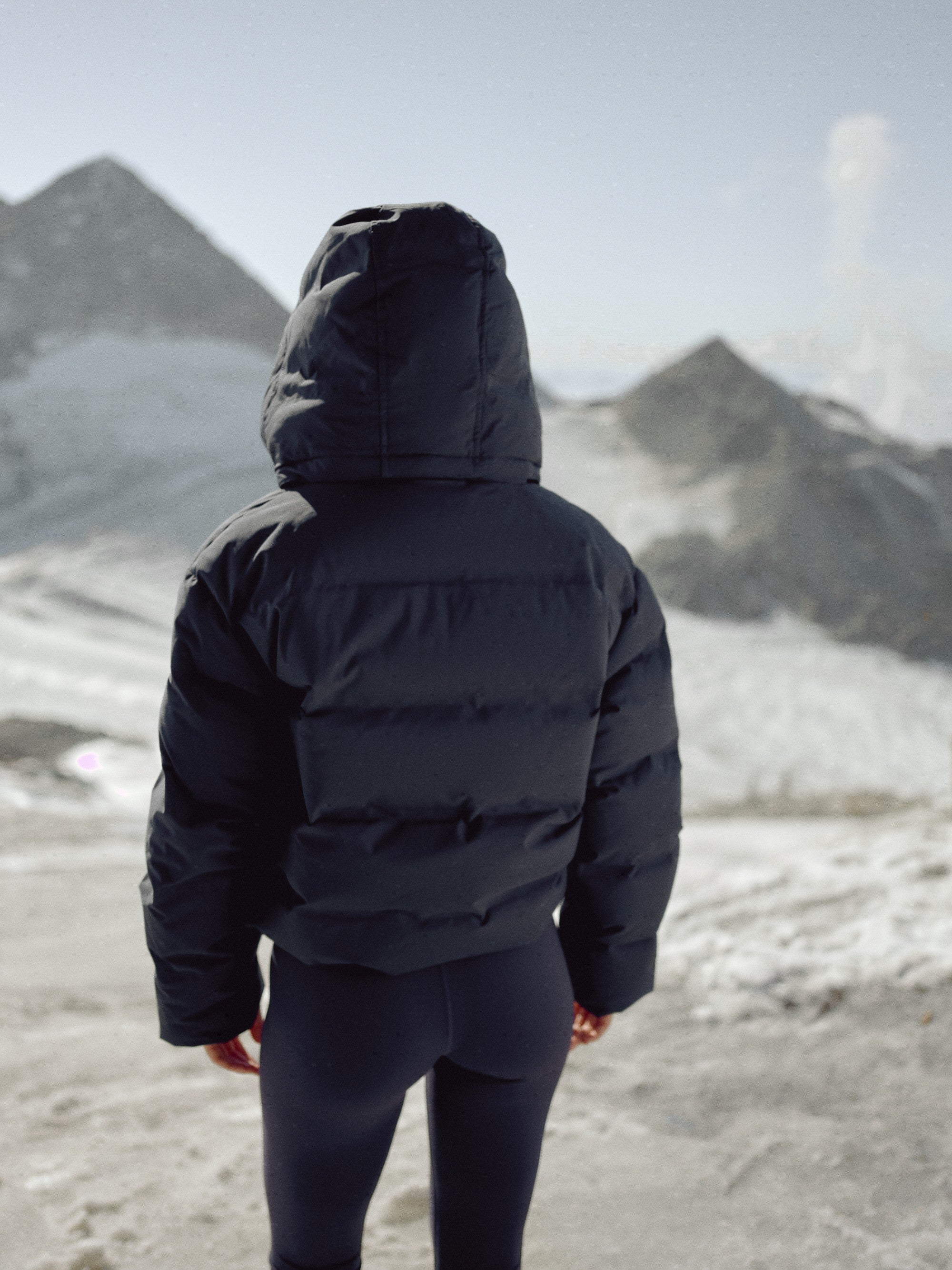 Back view of a woman wearing a navy CASPARA Aurora puffer jacket in the snowy mountains.