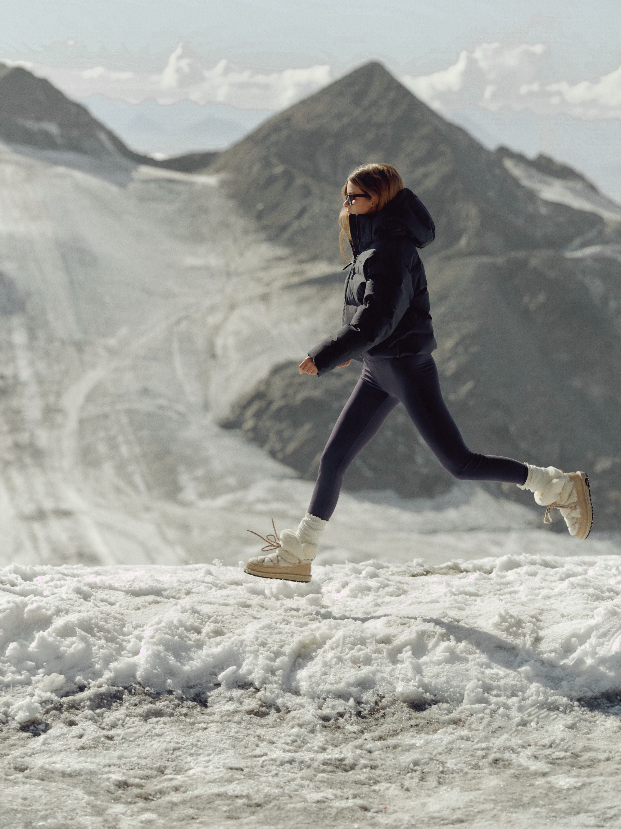 Woman leaping across snow wearing a navy CASPARA Aurora puffer jacket in the mountains.