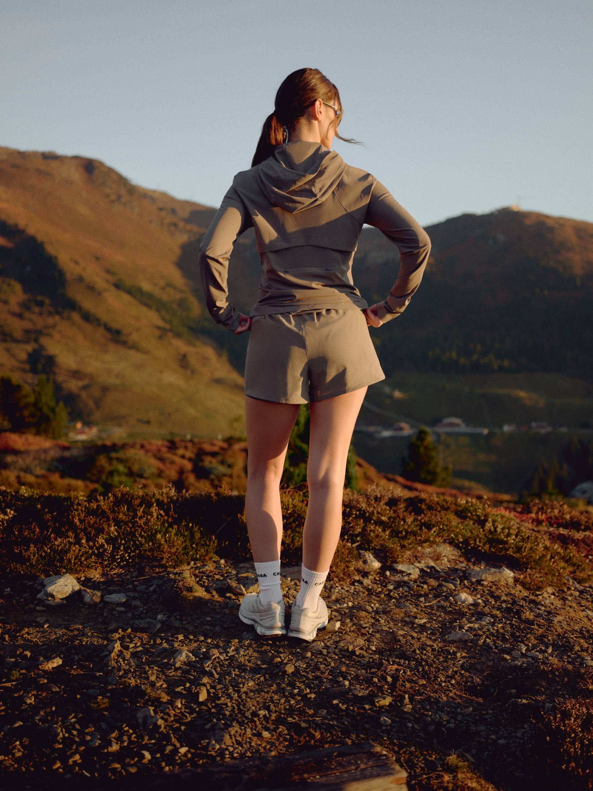 Woman wearing a khaki CASPARA Windluxe jacket standing on a mountain hillside.