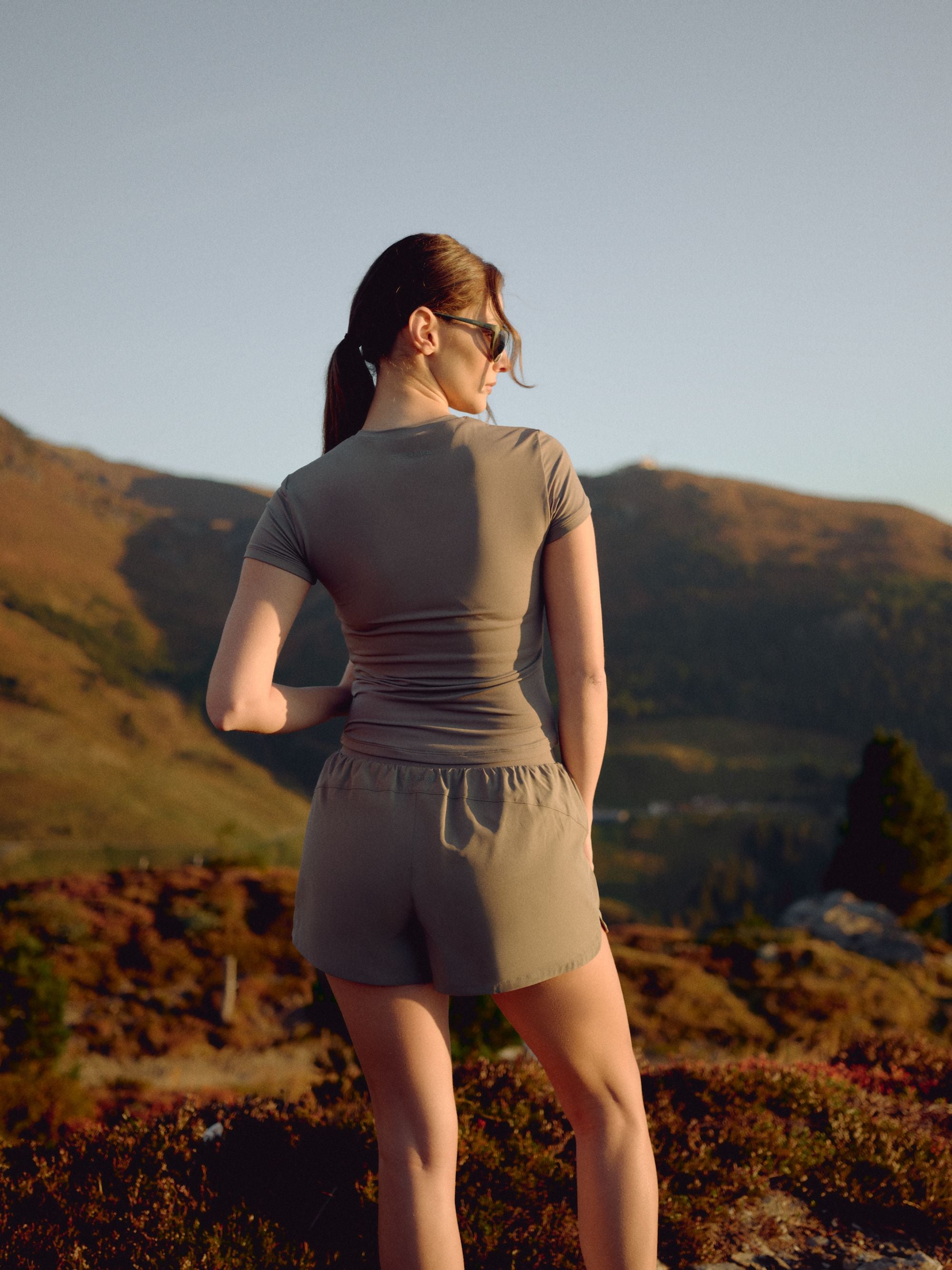 Woman wearing a khaki CASPARA activewear top and shorts in a mountain landscape.