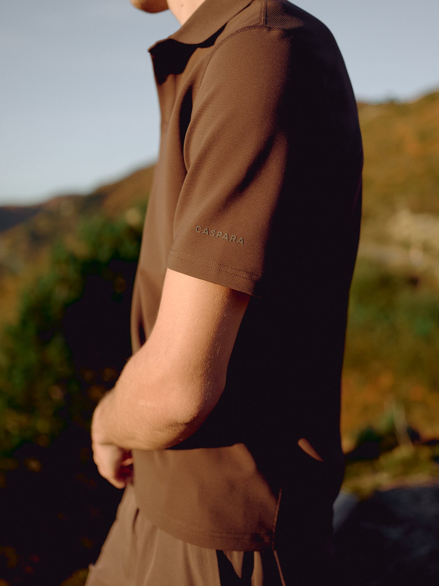 Side view of a man wearing an espresso CASPARA Vantage polo tee outdoors.
