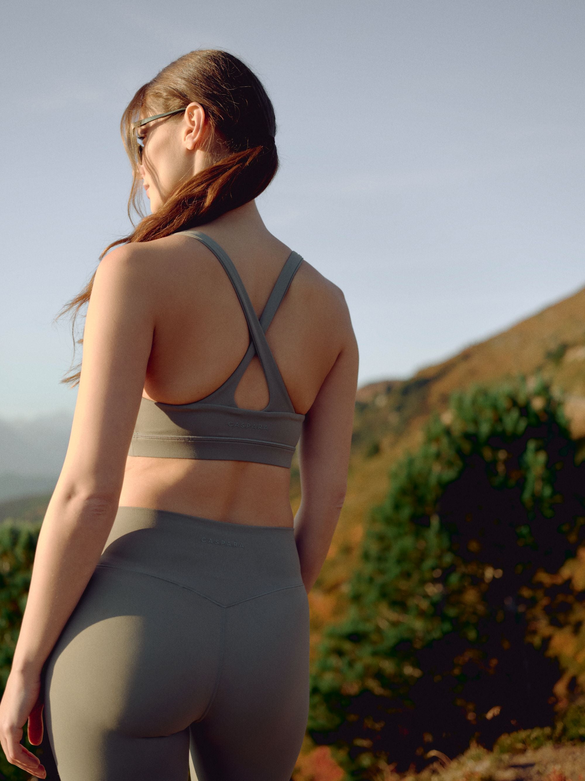 Woman wearing a khaki CASPARA Cloudform crossback sports bra on a sunny hillside.