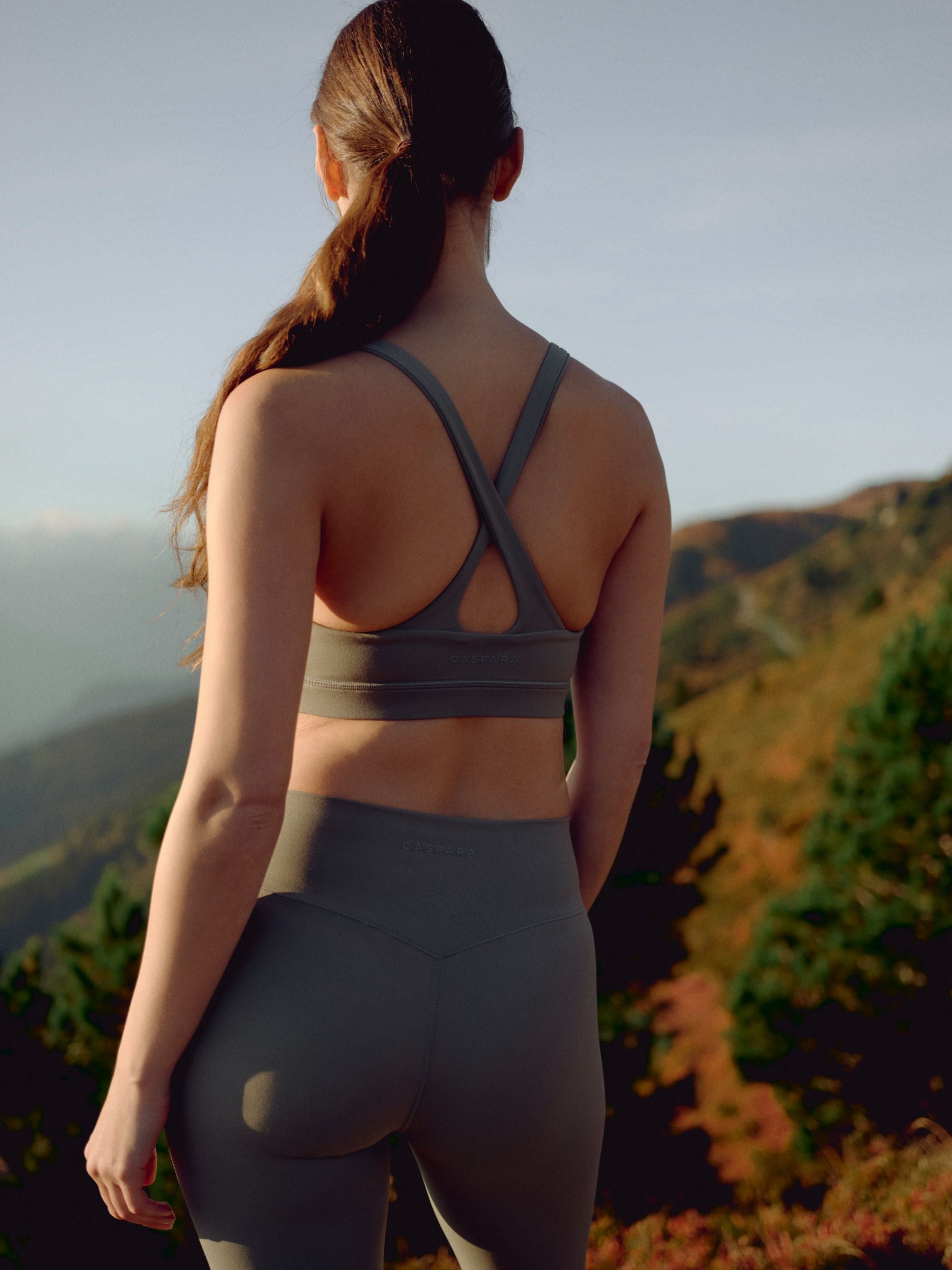Woman wearing a khaki CASPARA Cloudform crossback sports bra viewed from behind on a hillside.