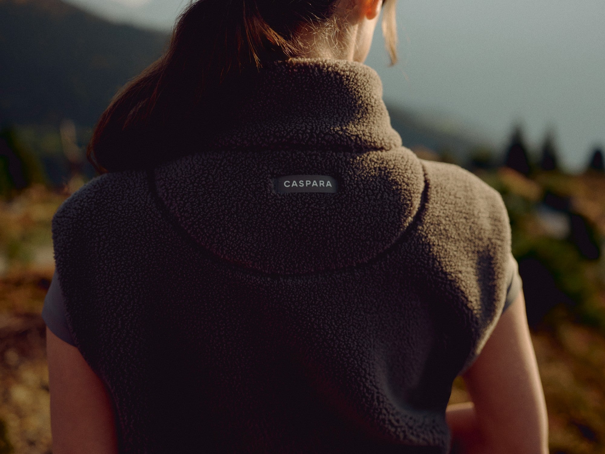 Back view of a woman wearing a khaki CASPARA Cloudfleece vest outdoors.