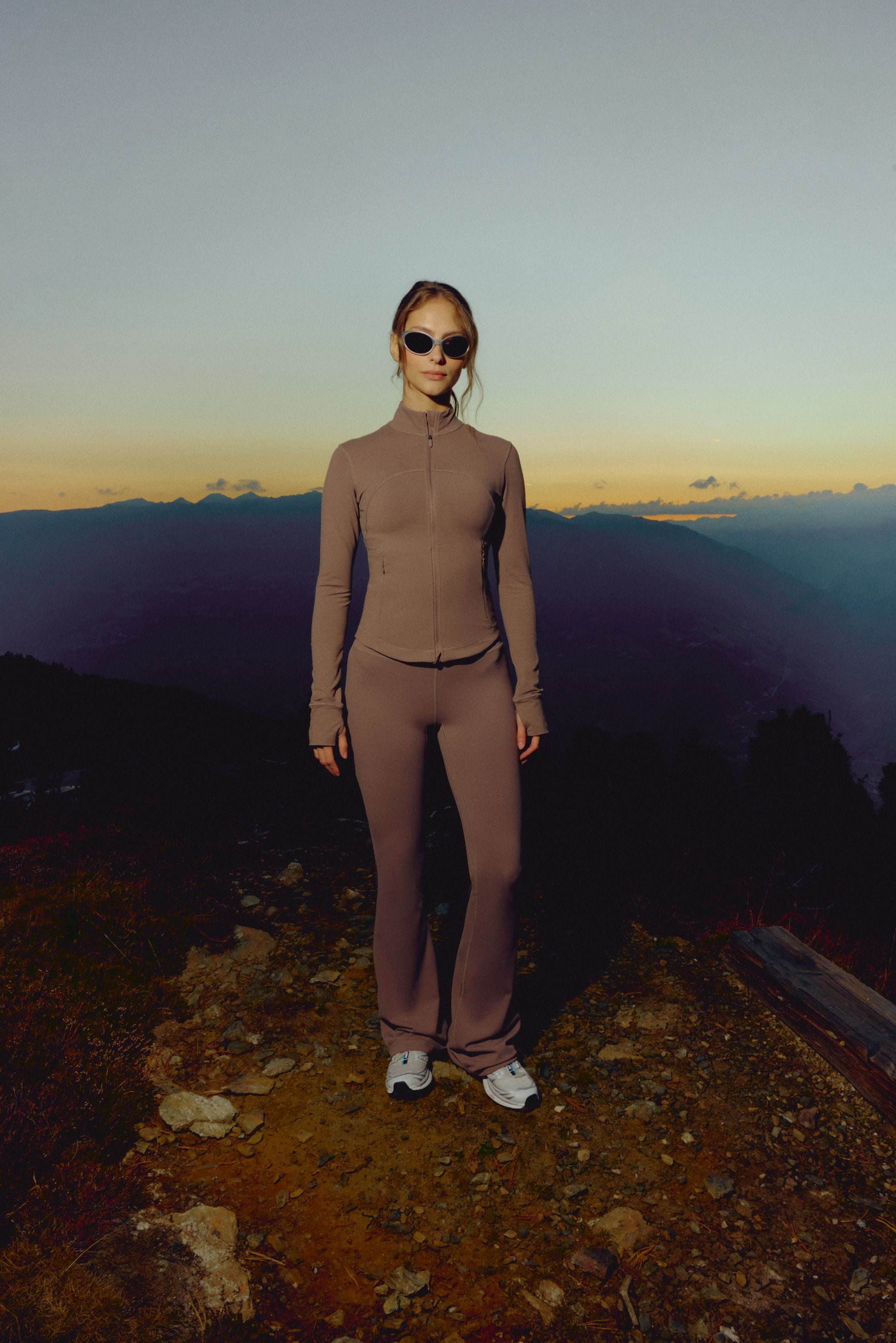 Woman wearing a walnut CASPARA Sculptluxe zip jacket standing on a mountain trail at sunset.
