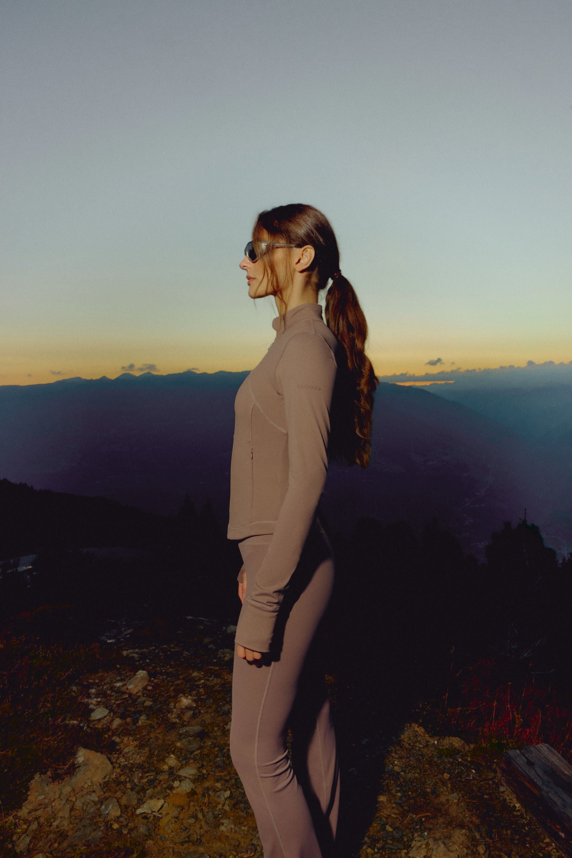 Woman wearing a walnut CASPARA Sculptluxe zip jacket standing on a mountain ridge at dusk.