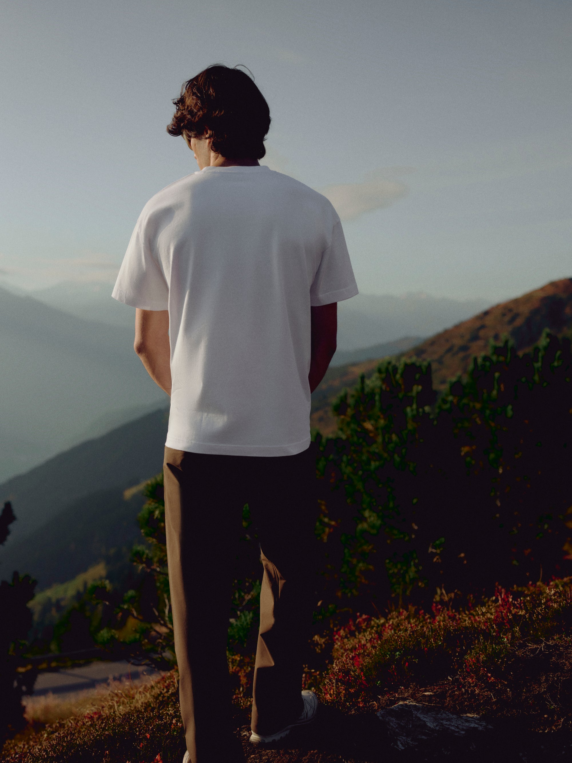 Back view of a man wearing a white CASPARA CS Signature t-shirt on a mountain hillside.