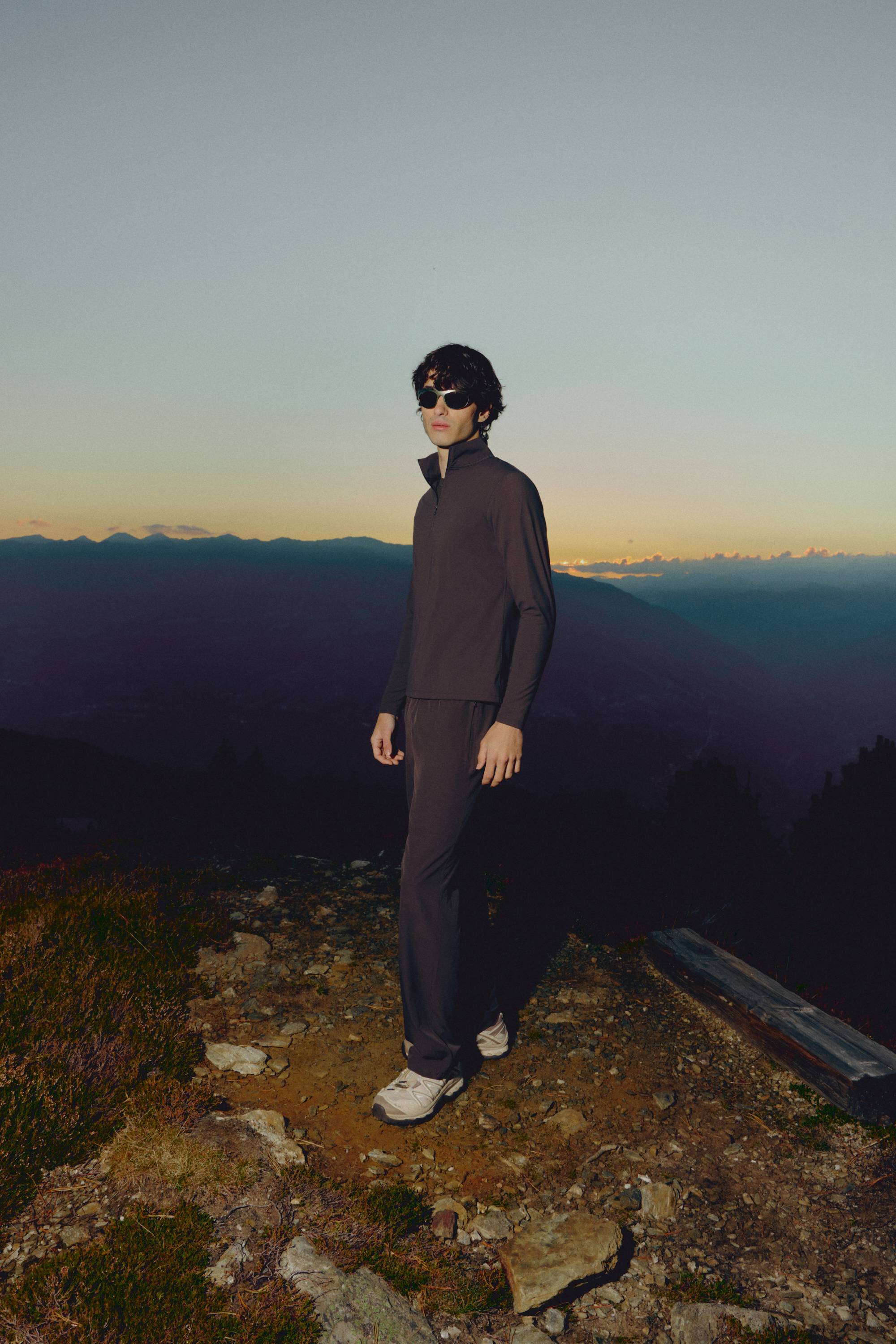 Man wearing espresso CASPARA Prime Core pants standing on a mountain hillside at dusk.