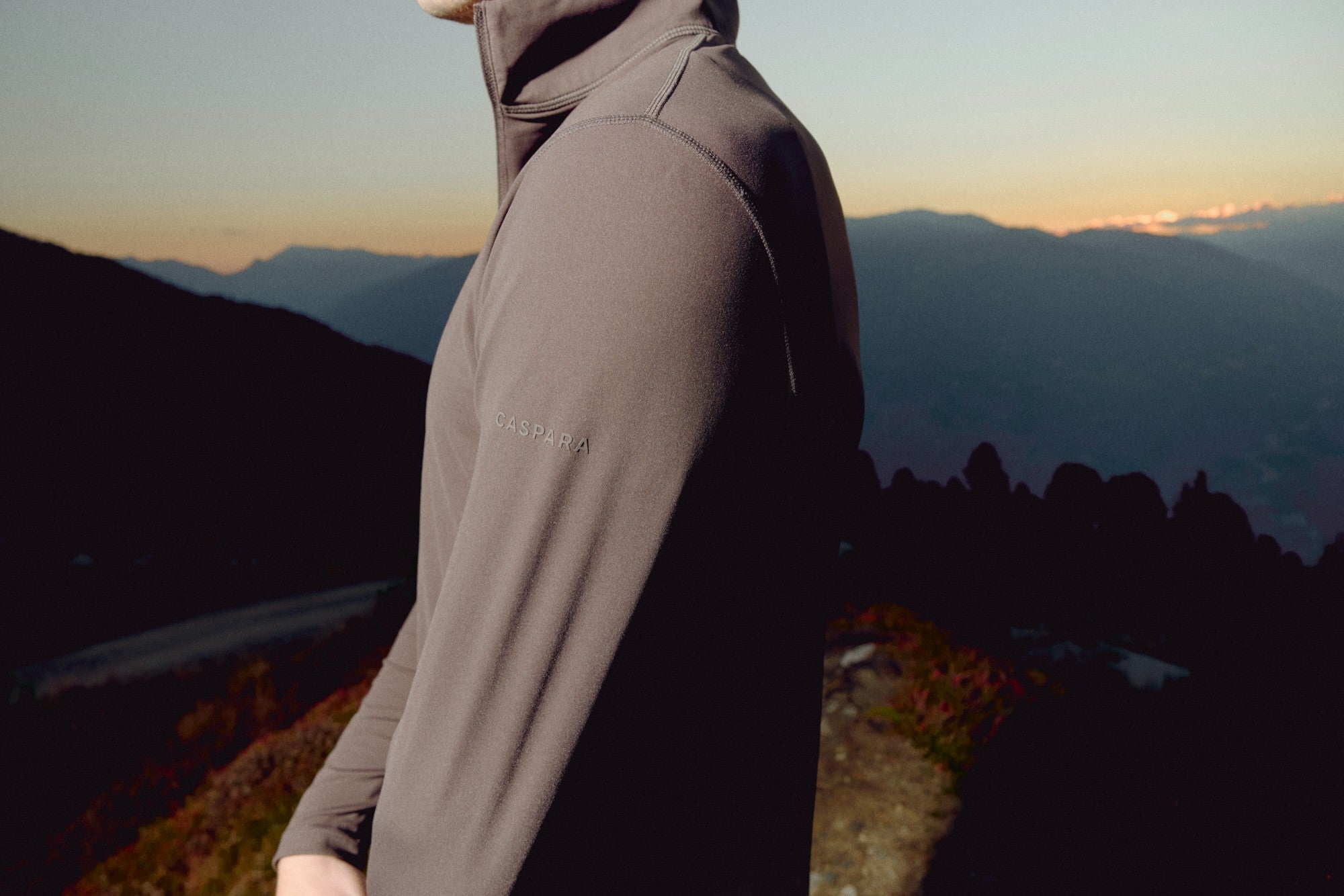Man wearing an espresso CASPARA Prime Core half zip overlooking a mountain landscape at sunset.