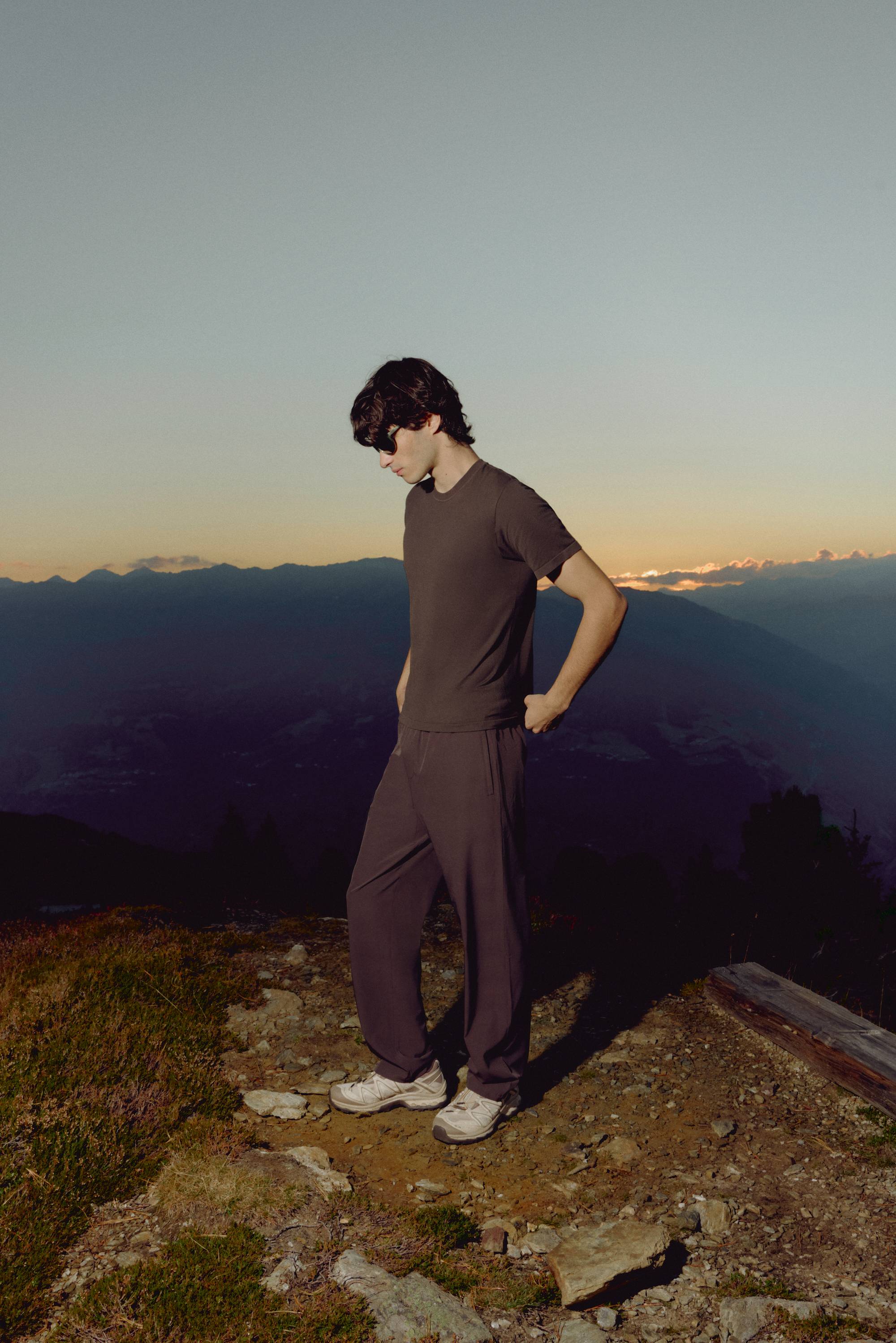 Man wearing an espresso CASPARA Seamlite t-shirt standing on a mountain ridge at sunset.