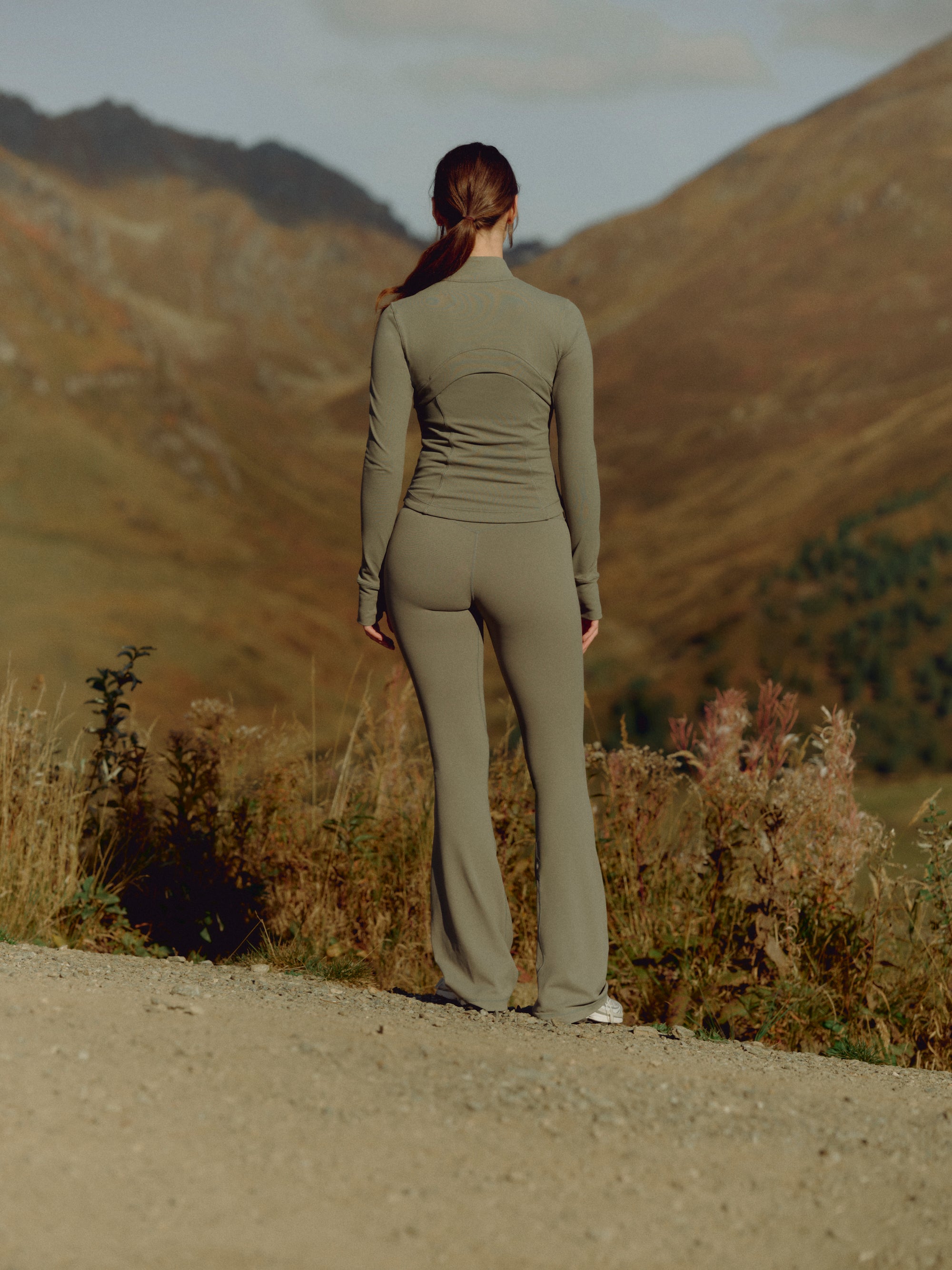 Woman seen from behind wearing a khaki CASPARA zip jacket and yoga pants in a mountain landscape.