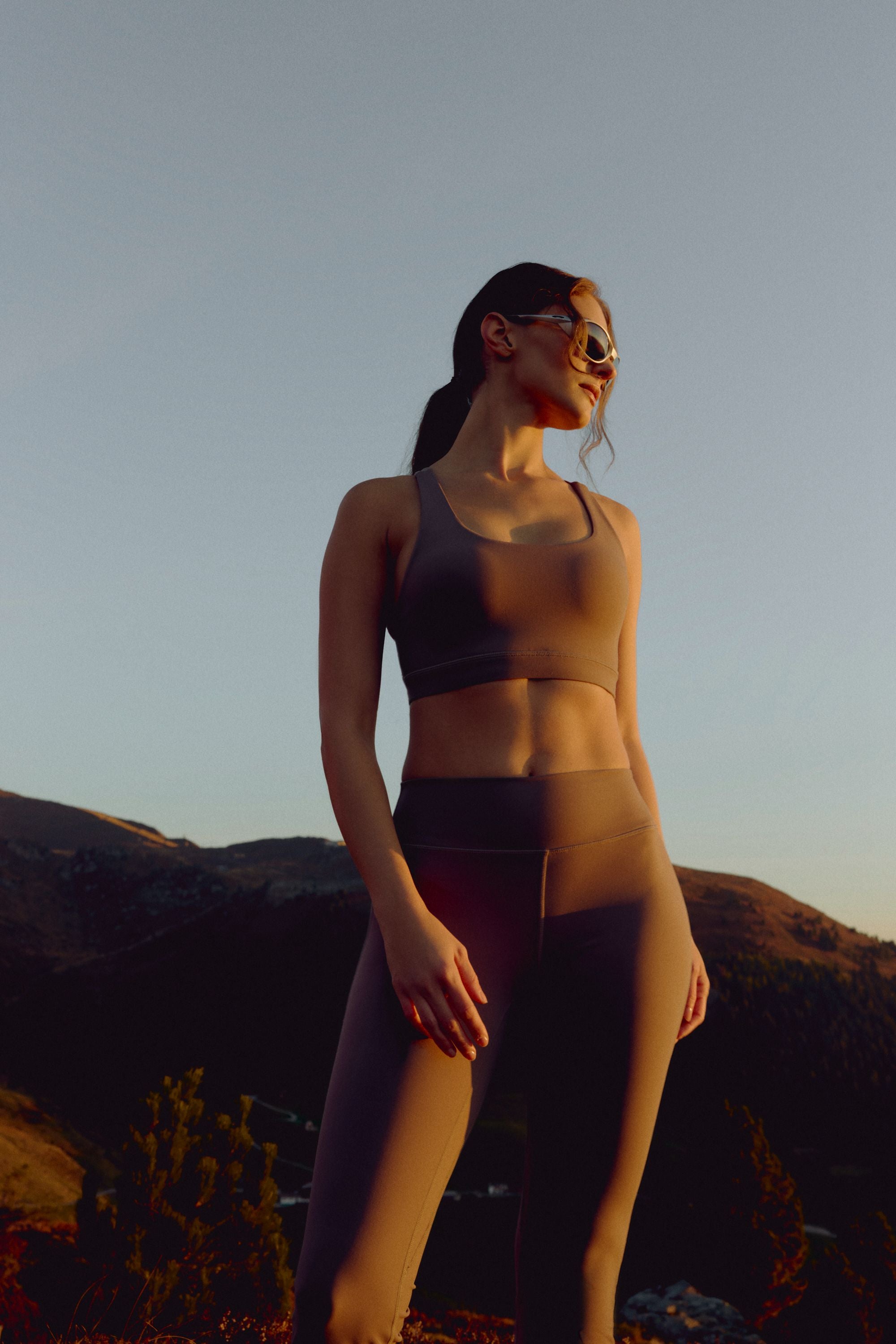 Woman wearing a walnut CASPARA Cloudform sports bra on a mountain hillside at sunset.