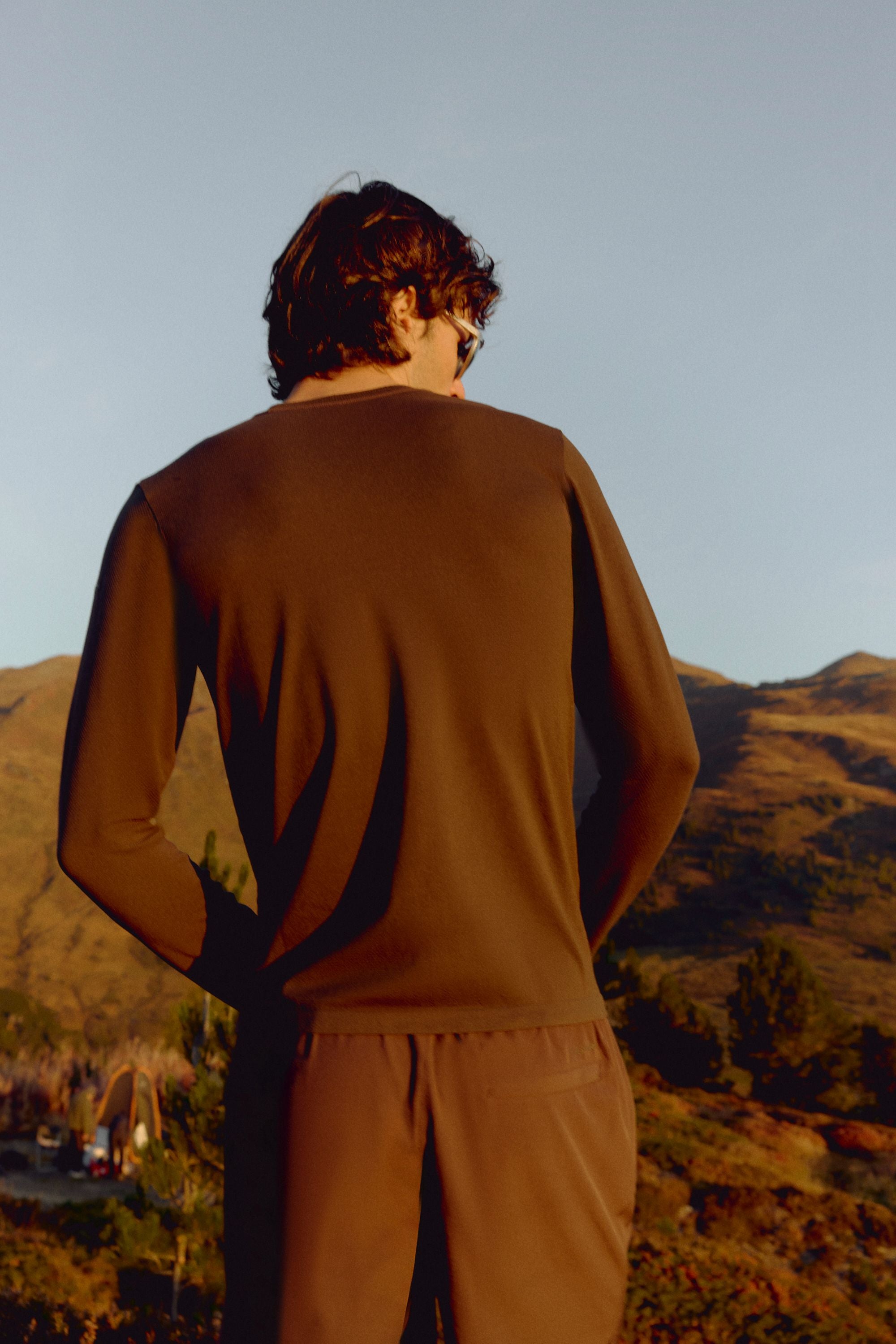 Back view of a man wearing an espresso CASPARA Seamlite long-sleeve shirt outdoors on a hillside.