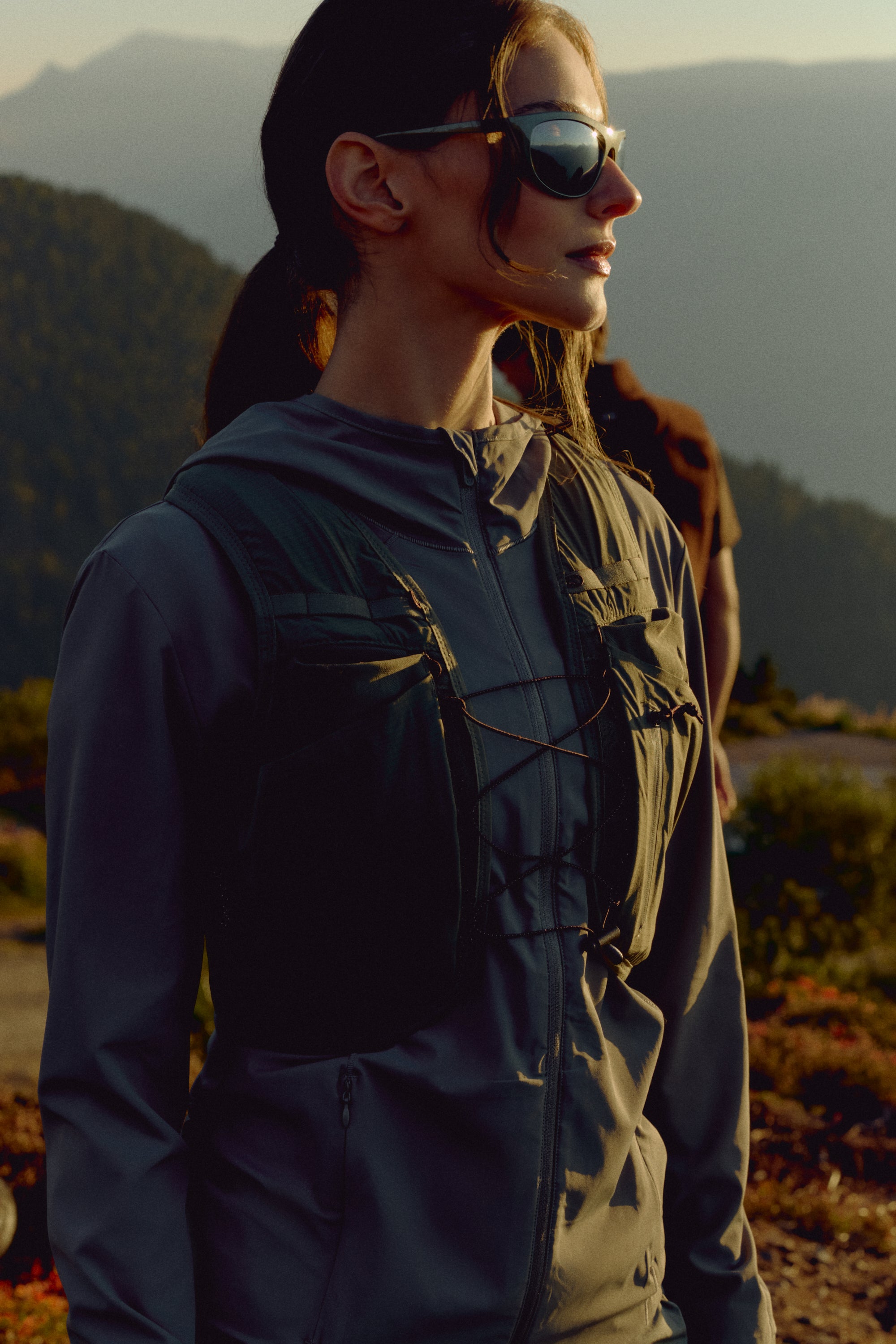 Woman wearing a khaki CASPARA Windluxe jacket and sunglasses on a mountain hillside.