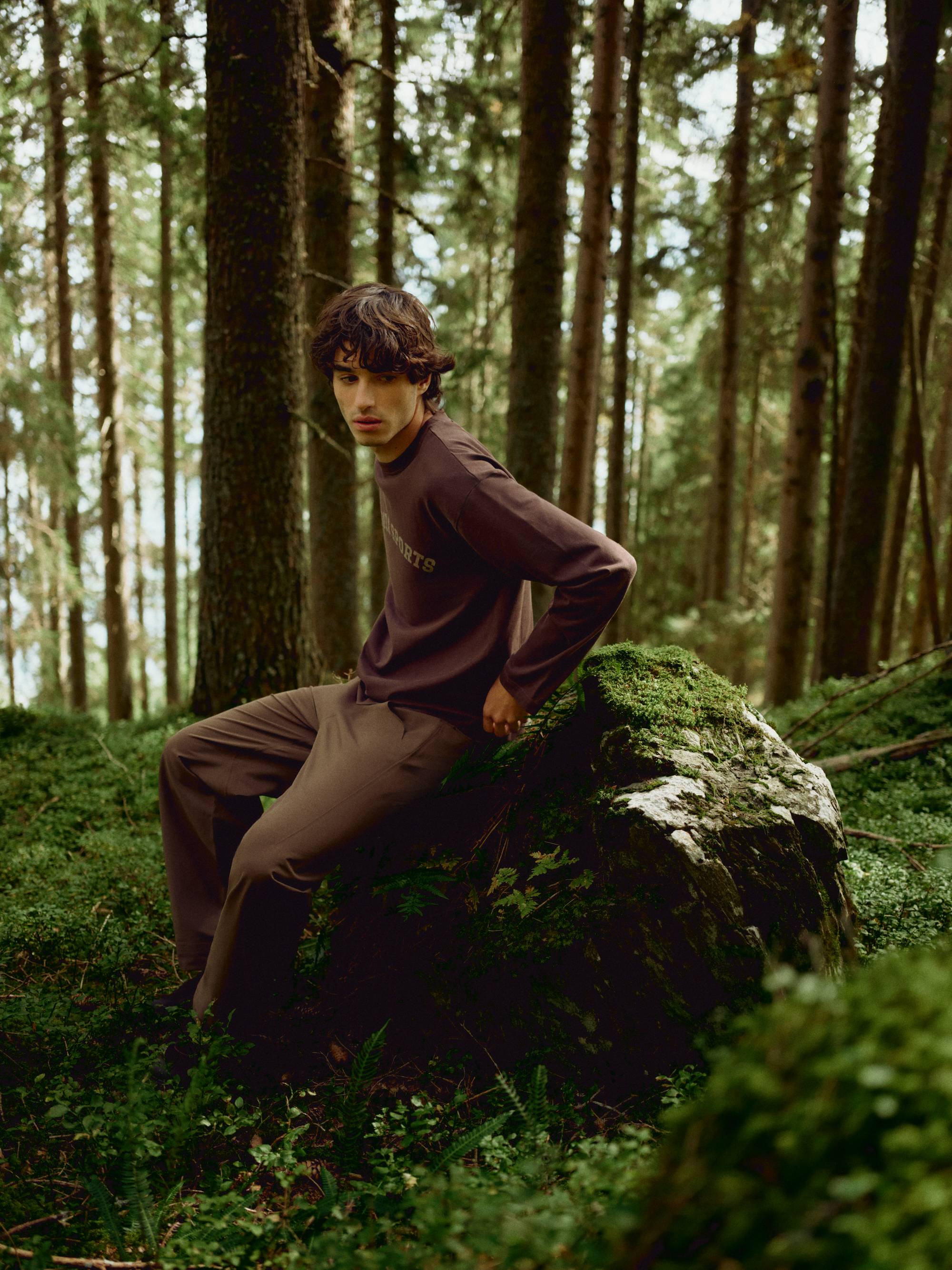 Man wearing an espresso CASPARA CS Signature long sleeve shirt sitting on a mossy rock in a forest.