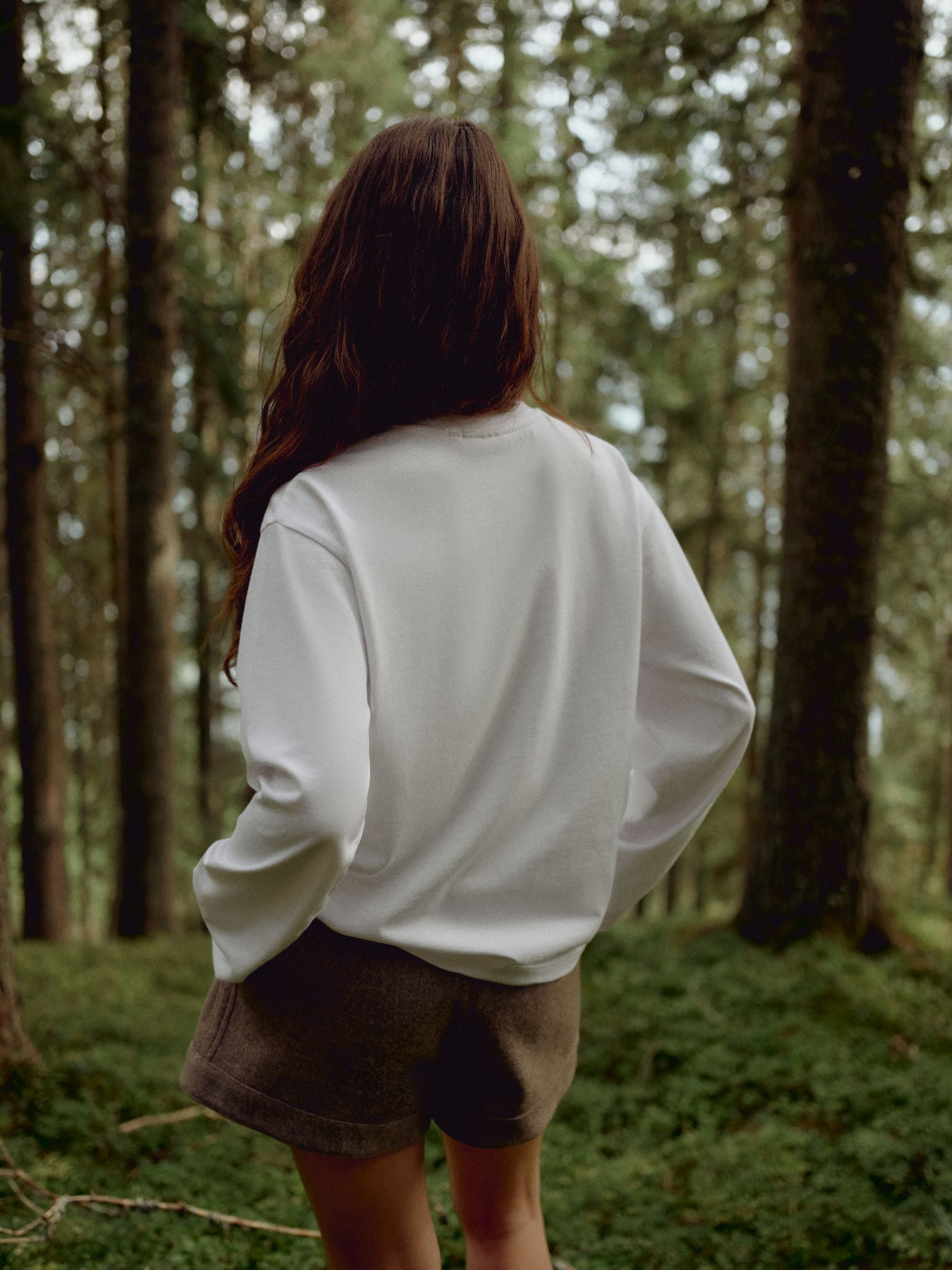 Woman wearing a white CASPARA CS Signature long sleeve shirt, seen from behind in a forest.