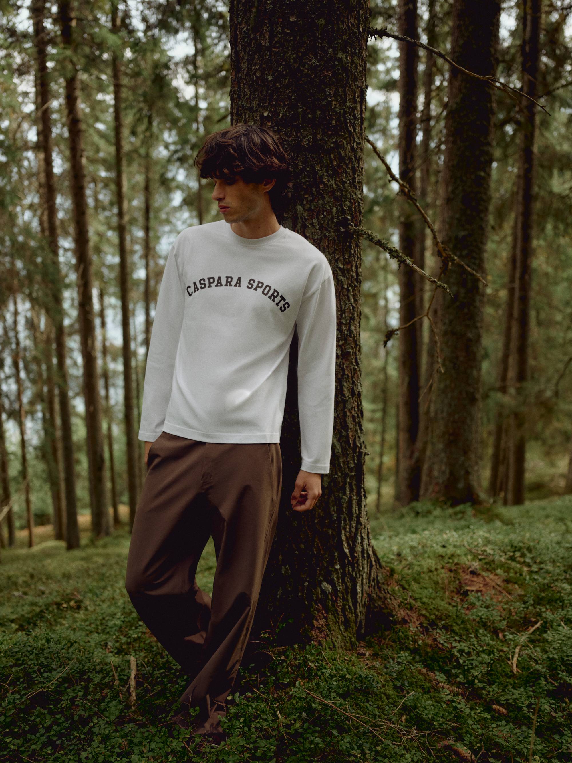 Man wearing a white CASPARA CS Signature long sleeve shirt leaning against a tree in a forest.