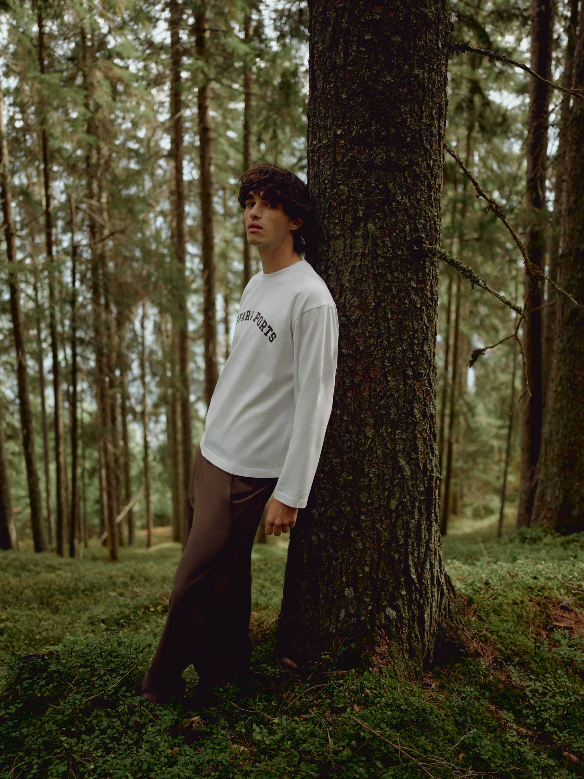 Man wearing a white CASPARA CS Signature long sleeve shirt leaning against a tree in a forest.