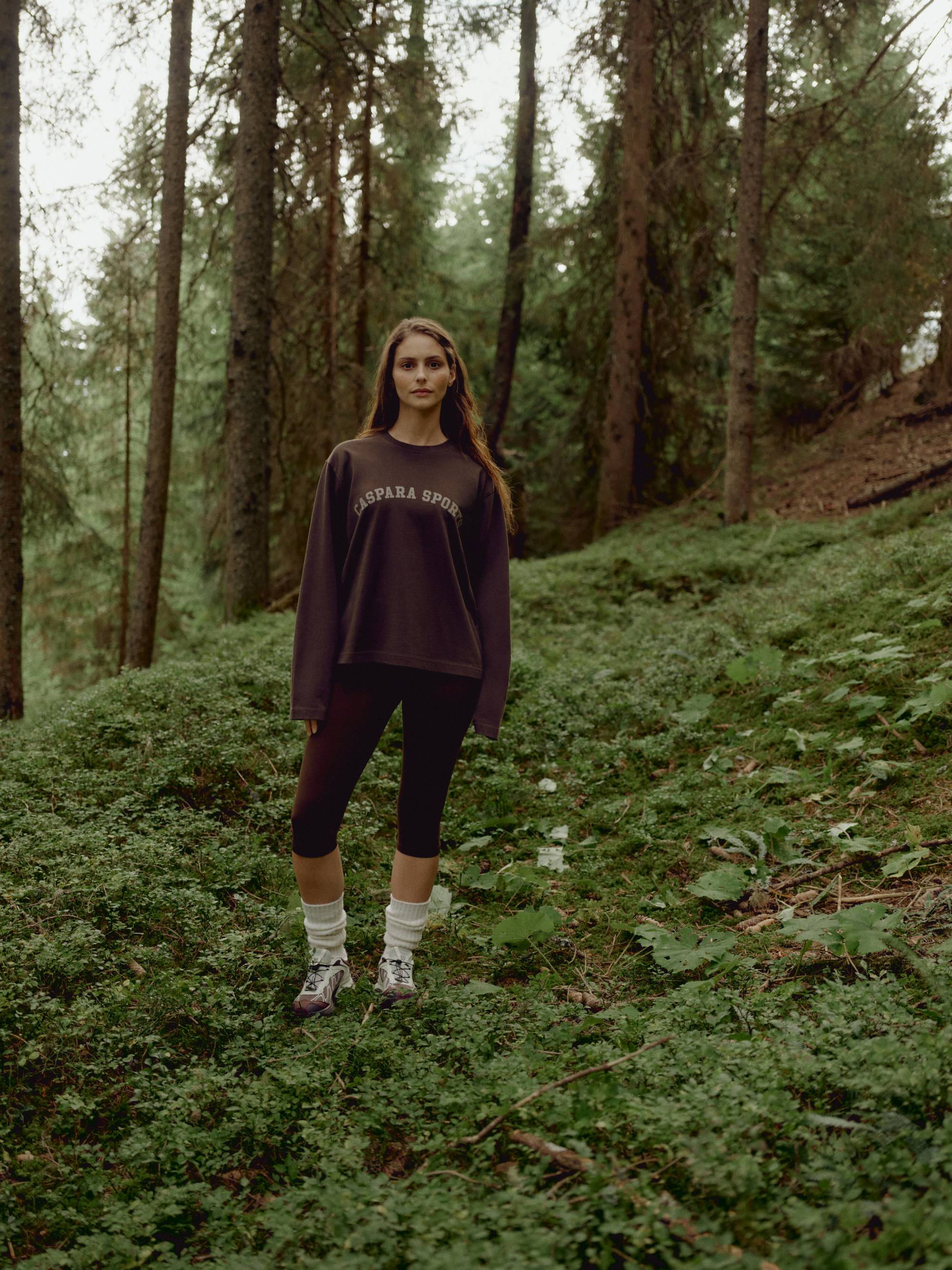 Woman wearing an espresso CASPARA CS Signature long sleeve shirt standing in a forest.