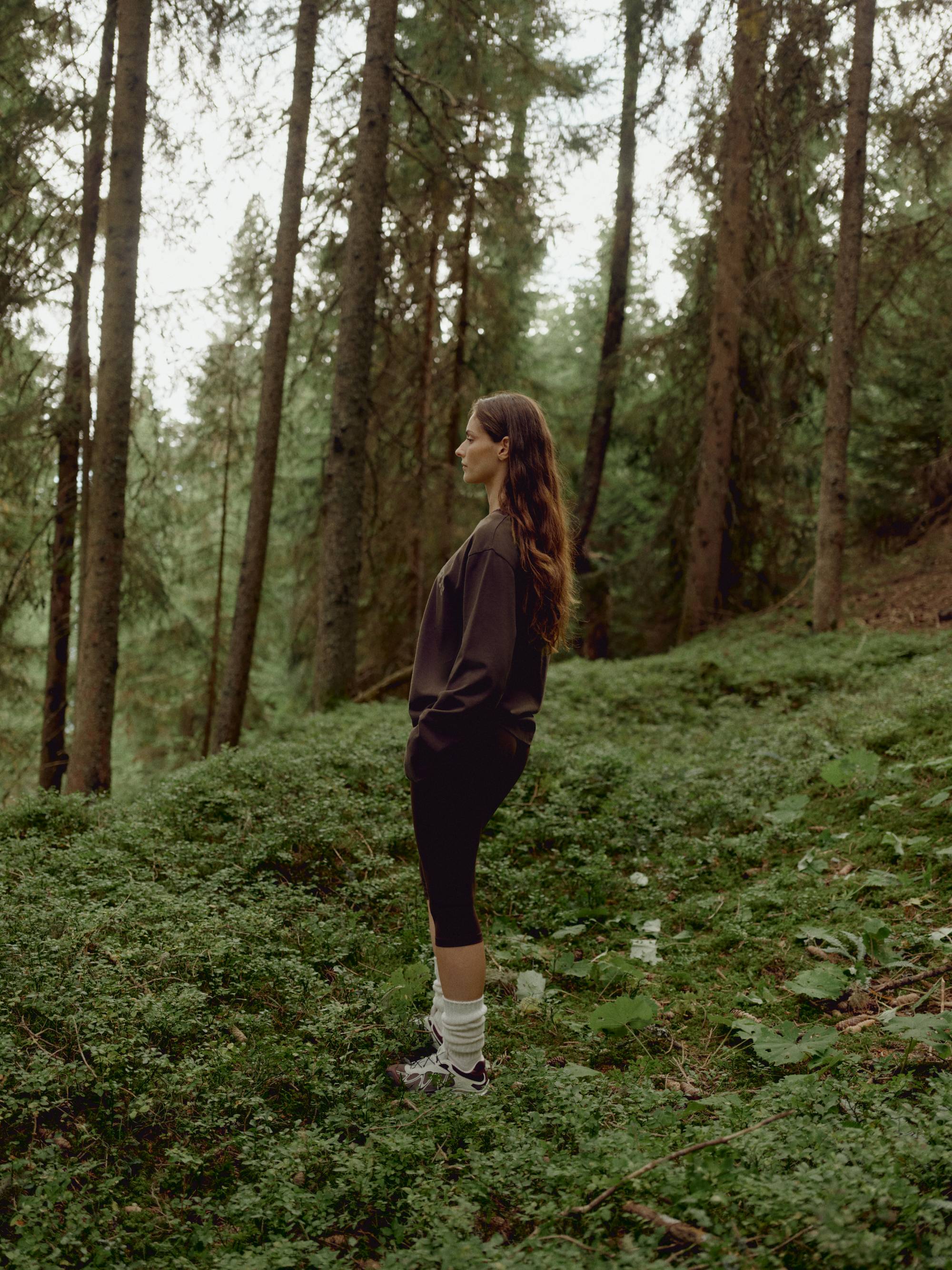 Woman wearing an espresso CASPARA CS Signature long sleeve shirt standing in a pine forest.