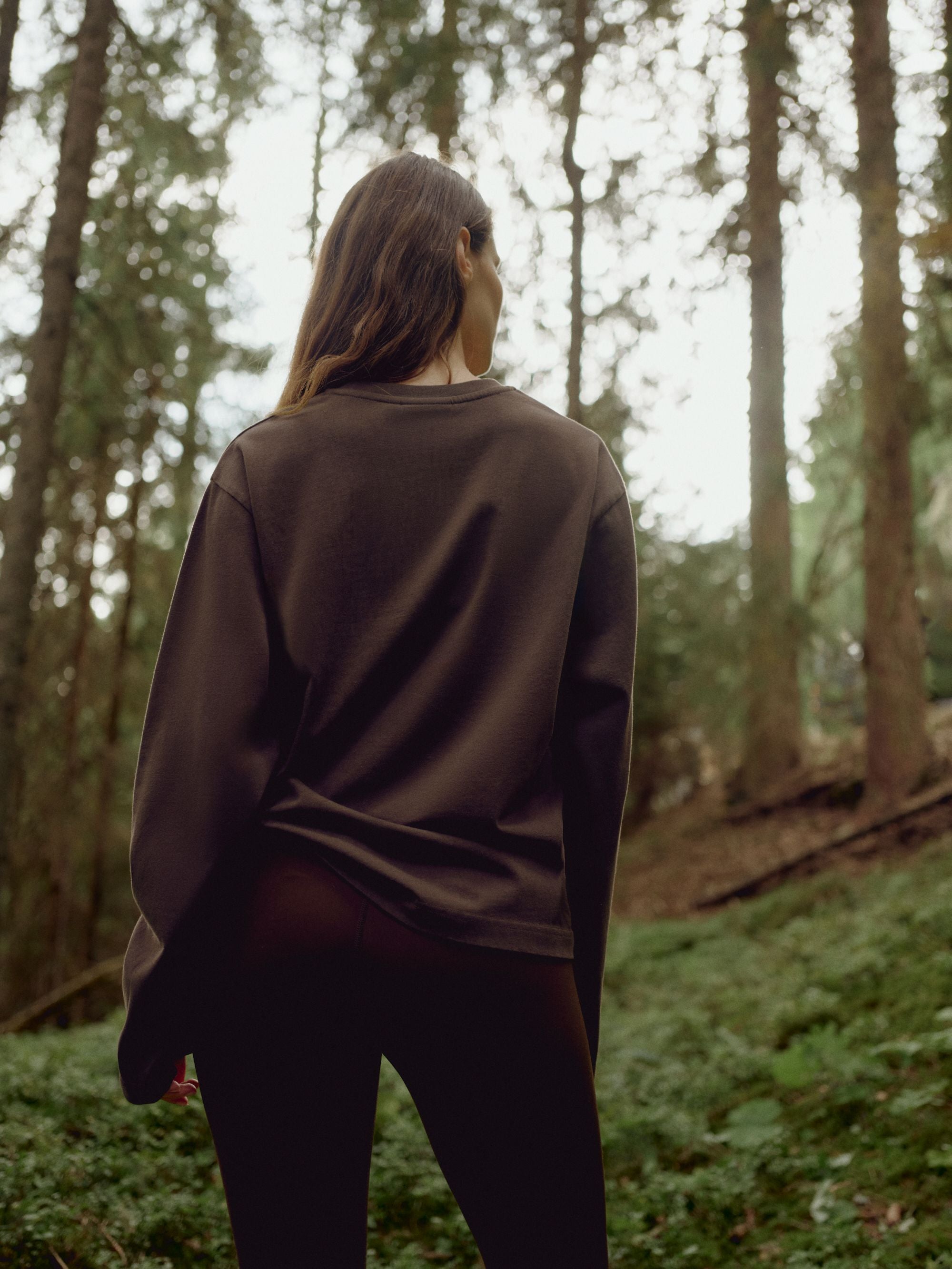 Woman wearing an espresso CASPARA CS Signature long sleeve shirt, seen from behind in a forest.