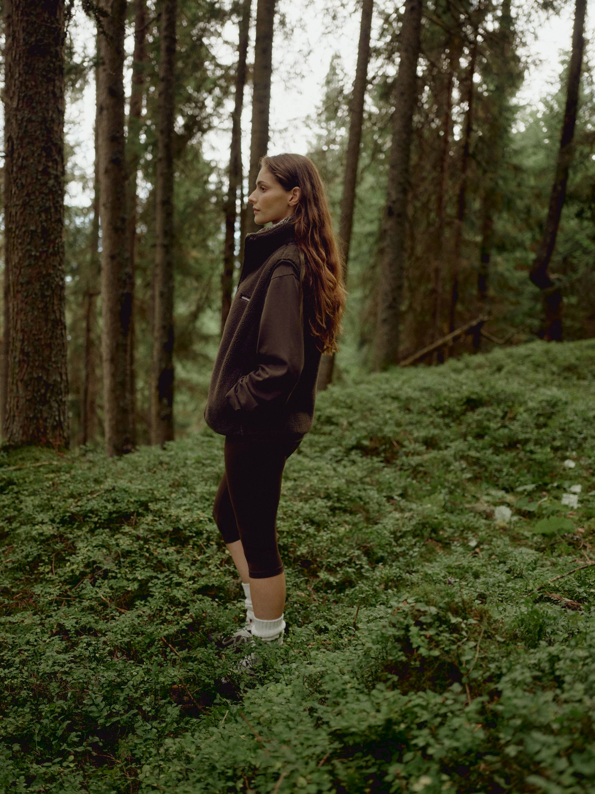 Woman wearing a khaki CASPARA Cloudfleece vest standing in a pine forest.