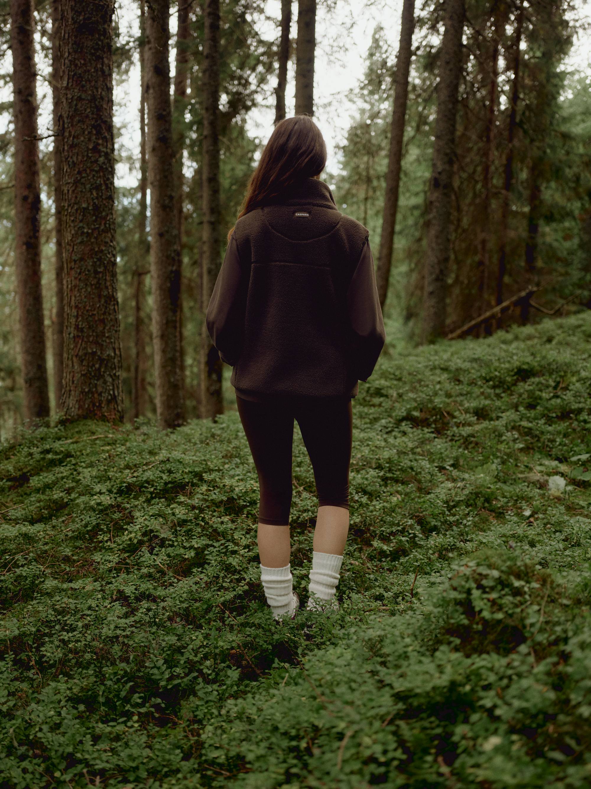 Back view of a woman wearing a khaki CASPARA Cloudfleece vest in a forest.