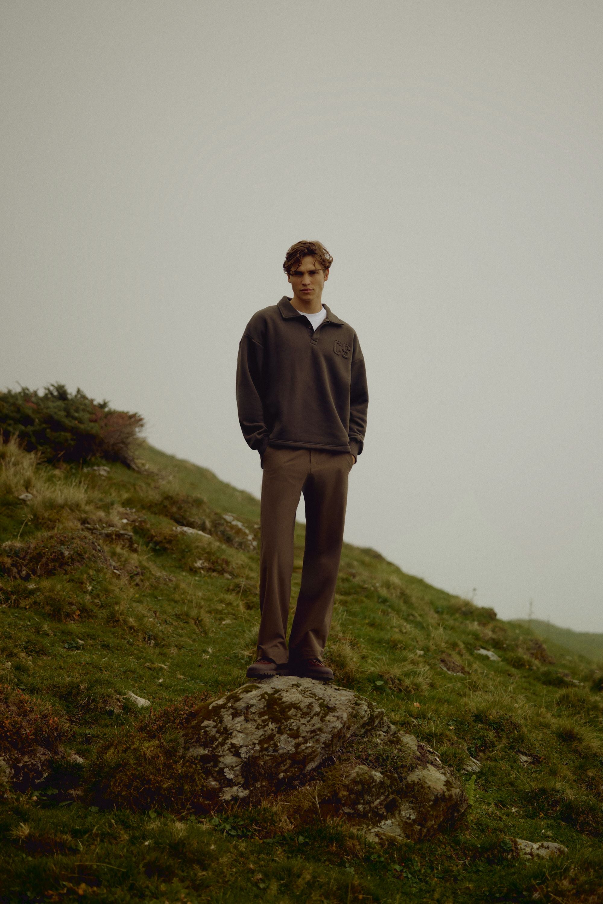 Man wearing an espresso CASPARA CS rugby shirt standing on a rocky hillside.