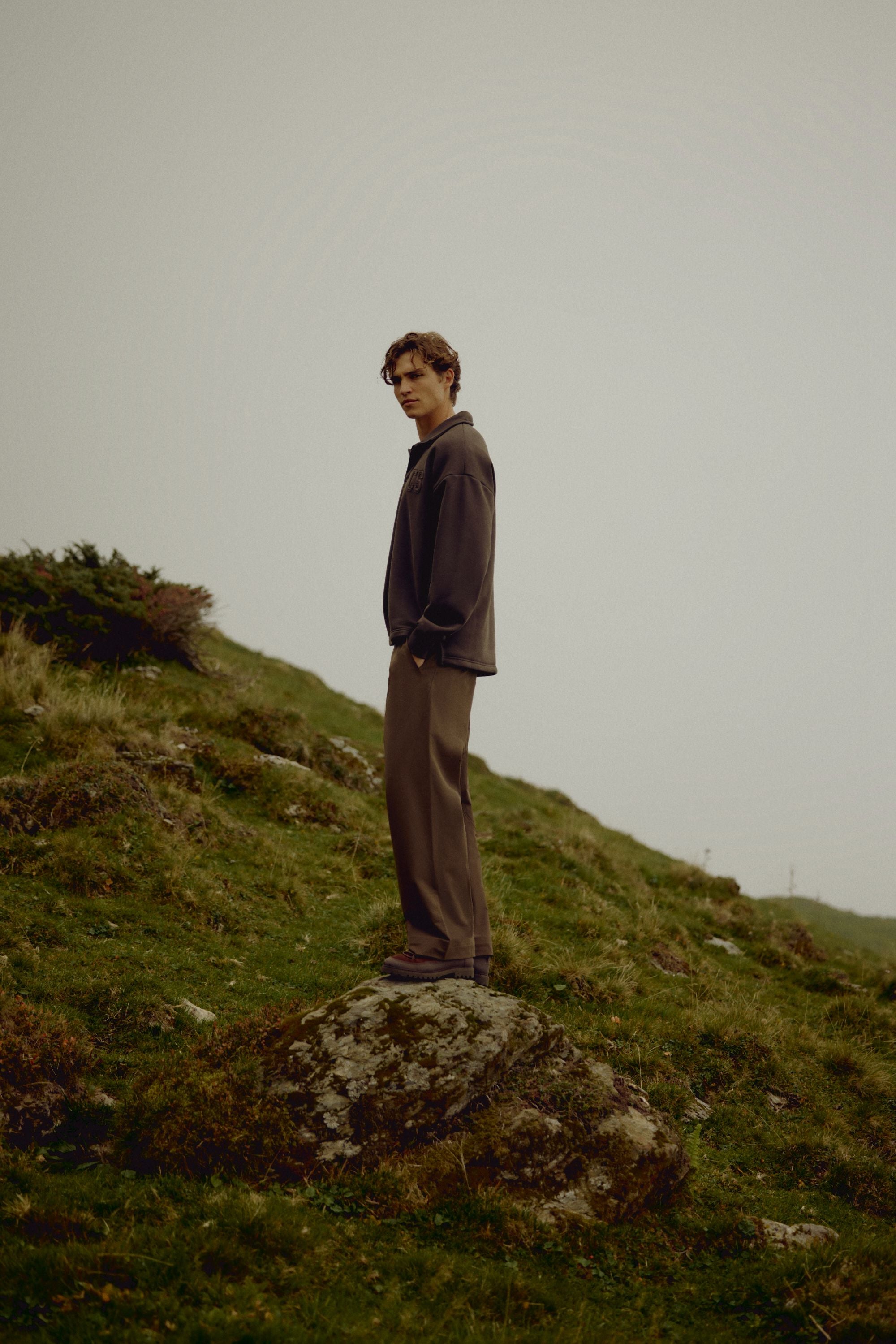 Man wearing an espresso CASPARA CS rugby shirt standing on a rocky hillside outdoors.